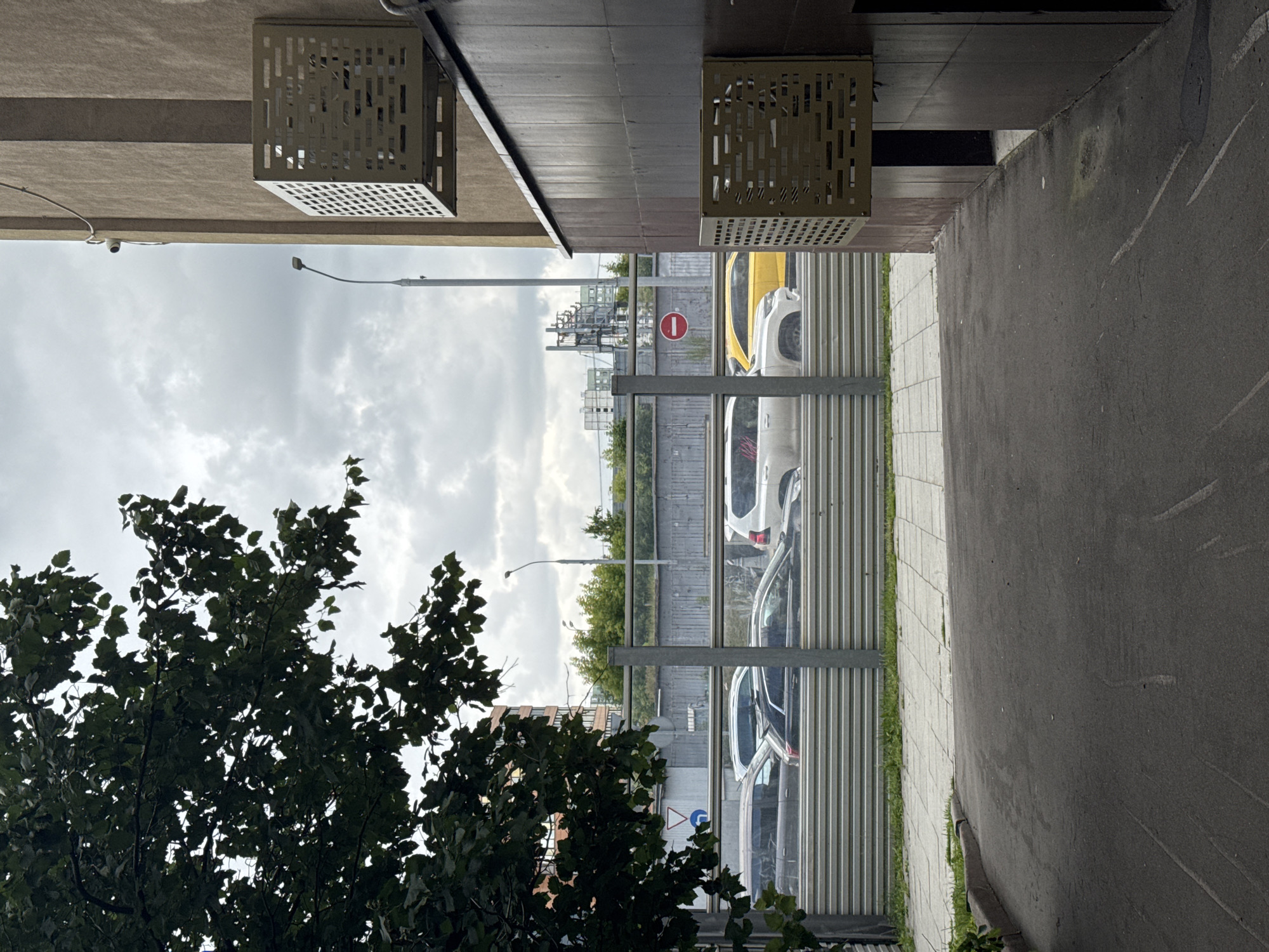 This image depicts an outdoor scene, likely in Moscow, Russia, under an overcast sky. The perspective is from underneath a building's overhang, looking outwards towards a street or parking area.
Dominating the foreground is a dark green, leafy tree, its branches obscuring parts of the view. Beyond the tree, a metal fence with corrugated panels separates the viewer from a row of parked cars. The cars appear to be a mix of colors, including white and yellow. One of the cars, a yellow hatchback, is particularly visible. Further in the background, there are signs of urban infrastructure, including what looks like a complex of industrial-looking pipes and structures, possibly a power station or factory. A red and white circular sign with a horizontal bar is also visible, indicating a restriction.
Above the street and cars, the sky is a muted gray, suggesting cloudy or possibly rainy weather. A street lamp with a curved arm is visible against the sky. The building's overhang features modern, geometric ventilation grates or decorative elements. The ground in the foreground is paved, with a strip of grass bordering the tiled walkway next to the parked cars.
The overall impression is a somewhat mundane urban environment, captured on a day with diffused light. There is no discernible activity or event taking place beyond the presence of parked vehicles. The image does not convey any strong emotions, appearing to be a straightforward depiction of a street-level view in a city. No legible text is visible in the image.