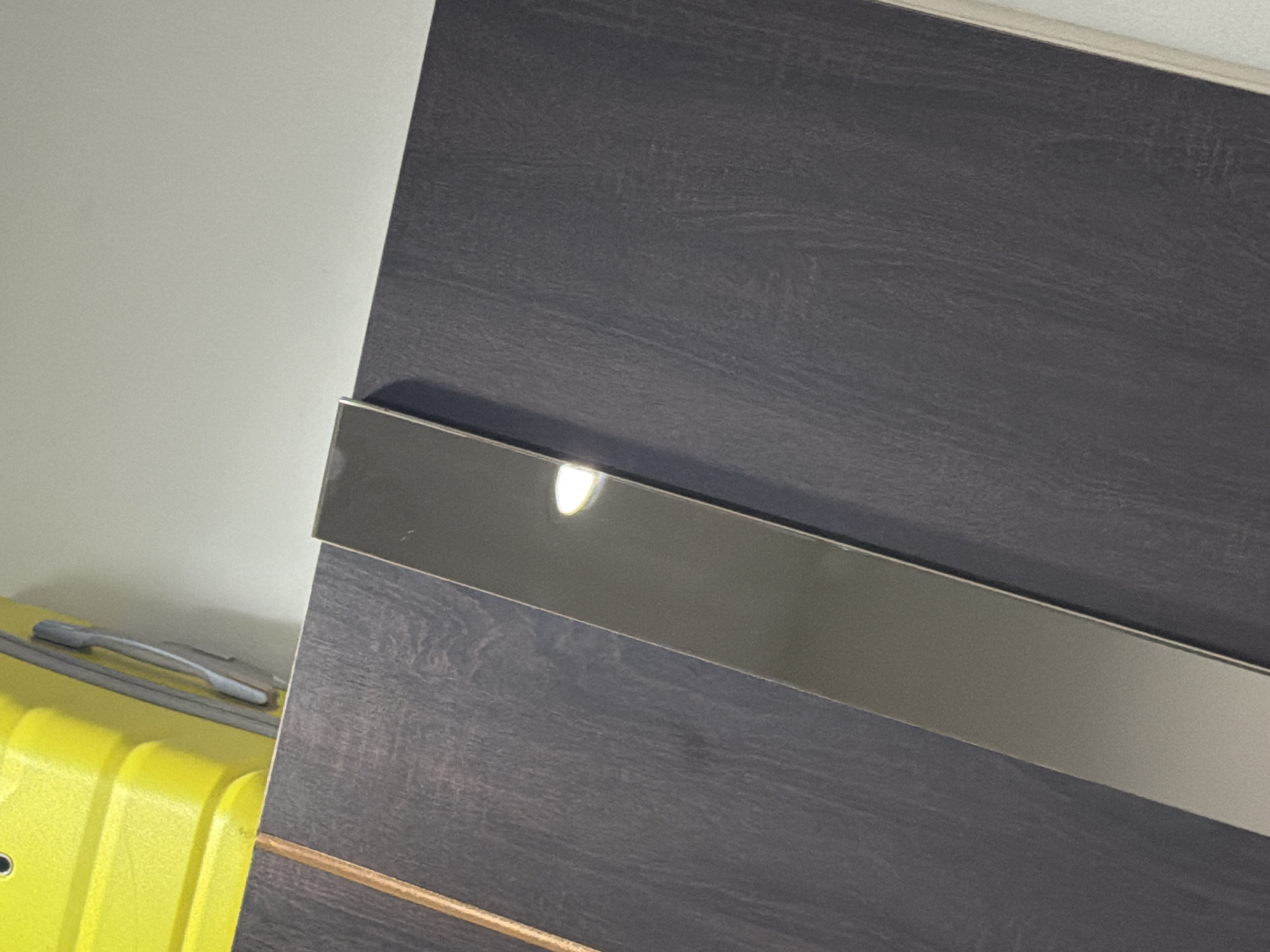 The image shows a close-up view of a dark gray wooden surface with a long, thin, reflective metallic strip running diagonally across it. A portion of a bright yellow object, possibly a piece of luggage, is visible in the upper left corner of the frame, suggesting an indoor environment. The background is a plain, light-colored wall. There are no people or animals present in the image, and no discernable activity is taking place. The scene appears to be a static composition focused on the textures and materials of the objects.