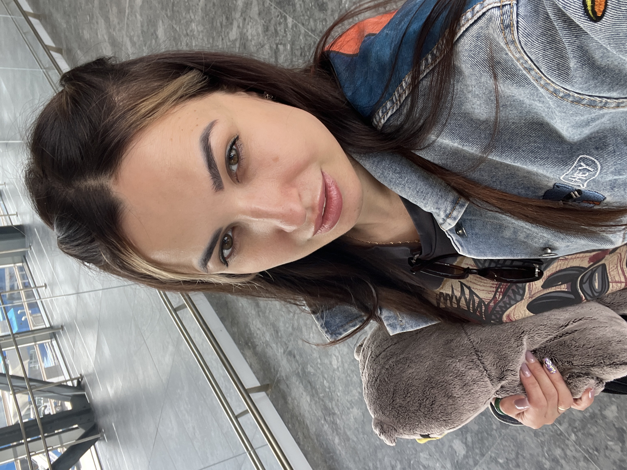 An eye-level, medium shot captures a young adult woman with long, dark brown hair styled in two small buns at the crown, holding a gray plush toy with both hands. She is wearing a denim jacket with colorful patches on the sleeves and a dark t-shirt underneath. Her face is turned towards the camera, and she has light makeup on. Sunglasses are hanging on her t-shirt. She is standing in what appears to be a modern public space with gray tiled flooring and a sleek metal railing. The background shows a blurred view of a walkway with large windows and structural elements, suggesting an indoor or covered outdoor area. The lighting is bright and natural.