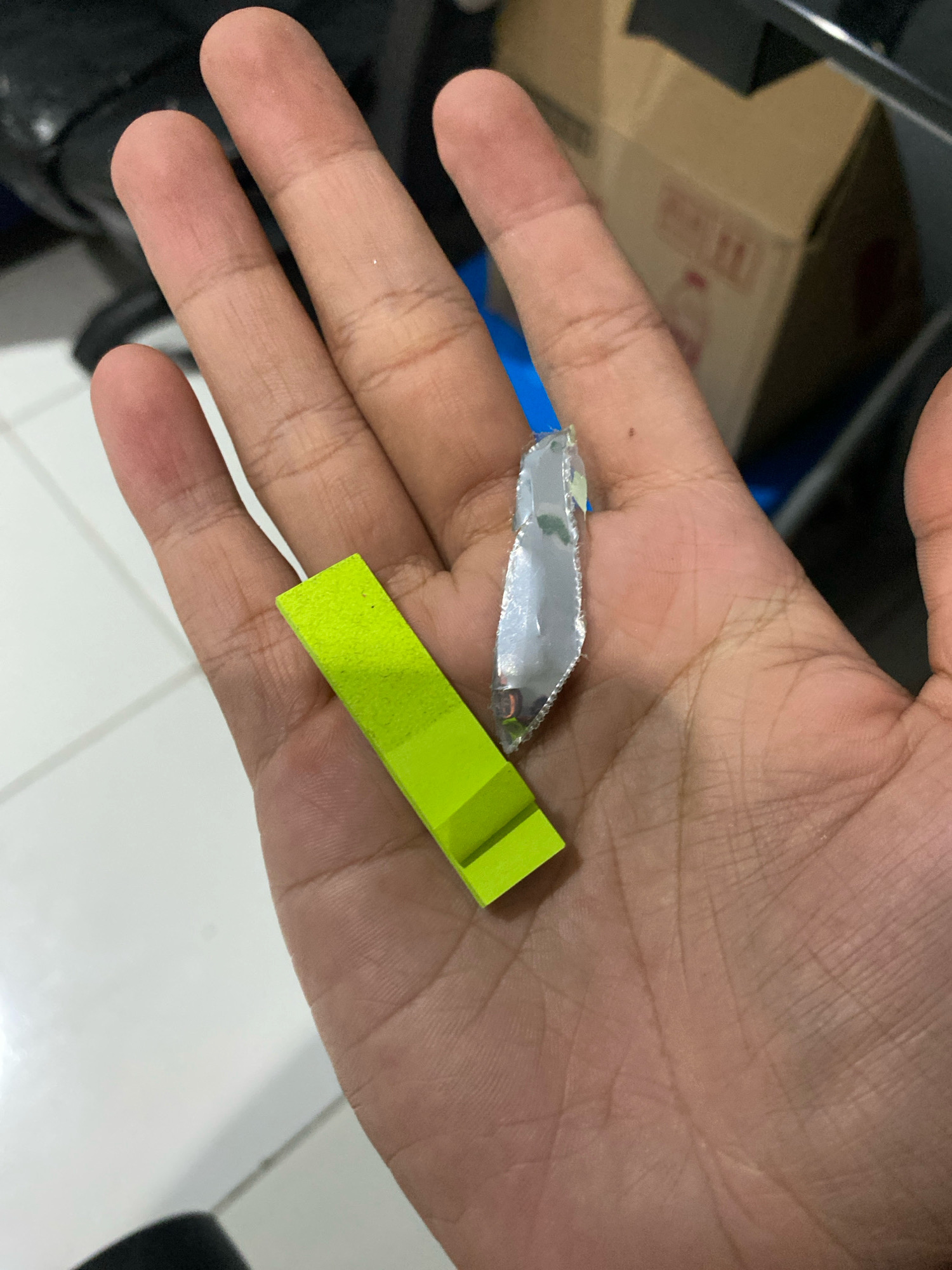 A close-up, top-down shot shows a person's open palm. Resting on the palm are two small objects. On the left, a bright lime green, rectangular prism-shaped object. To the right of this, a sliver of reflective, possibly metallic, material with jagged edges. The background is blurred, but appears to be an indoor setting with a white tiled floor and hints of furniture. The image does not depict any specific location within the city of Klurak, Indonesia, or any urban elements.