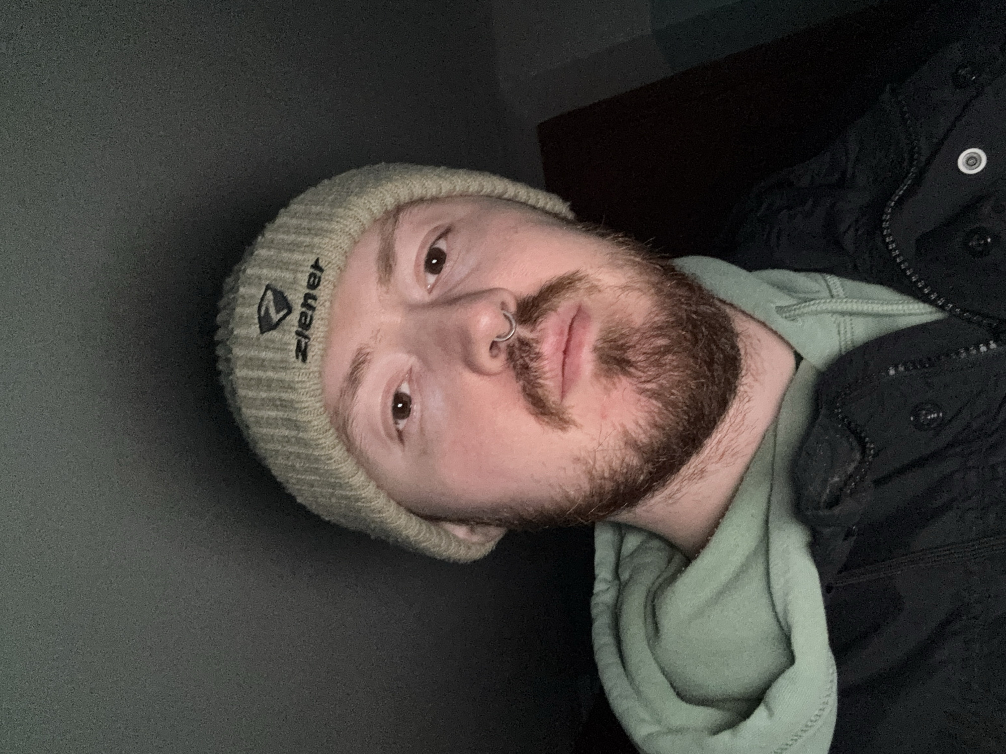 A man is depicted in an indoor setting. He is wearing a light-colored beanie with a logo on it, a light green hooded sweatshirt, and a black jacket. He has a beard and a nose ring. The background is dark, with a dark grey wall visible on one side. The location is not specified.