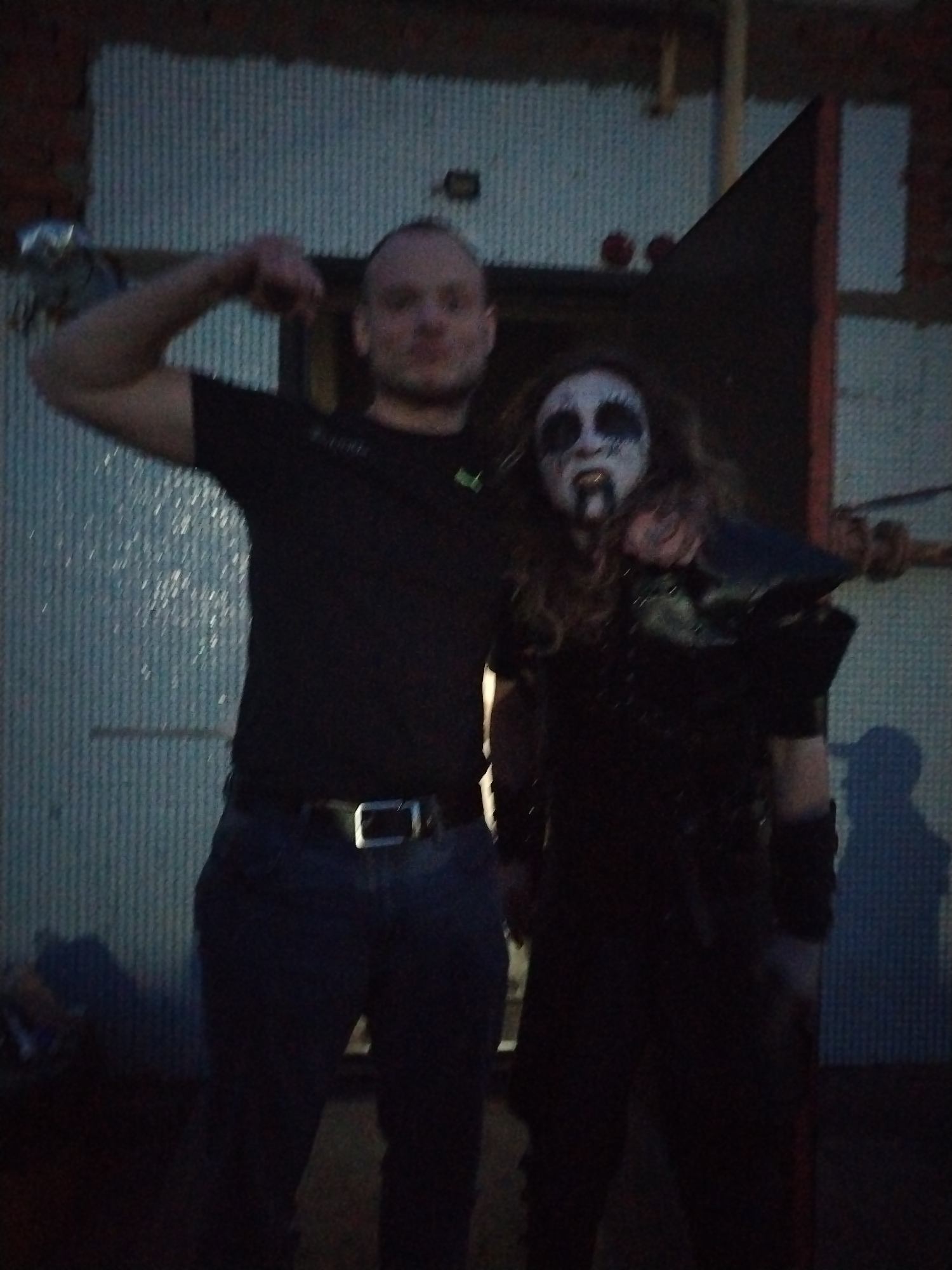 The image shows two people standing next to each other in an outdoor setting.
One person on the left is wearing a dark t-shirt and blue jeans with a belt. He is flexing his arm.
The second person on the right has dark hair and is wearing black clothing with a studded shoulder pad. They have pale face paint with black markings around their eyes and mouth.
The background features a corrugated metal wall and what appears to be a doorway or opening with red trim. There is also a brick wall visible in the background.
There are no specific details indicating the location is in Chaikovsky, Russia, other than the general appearance of the architectural style.