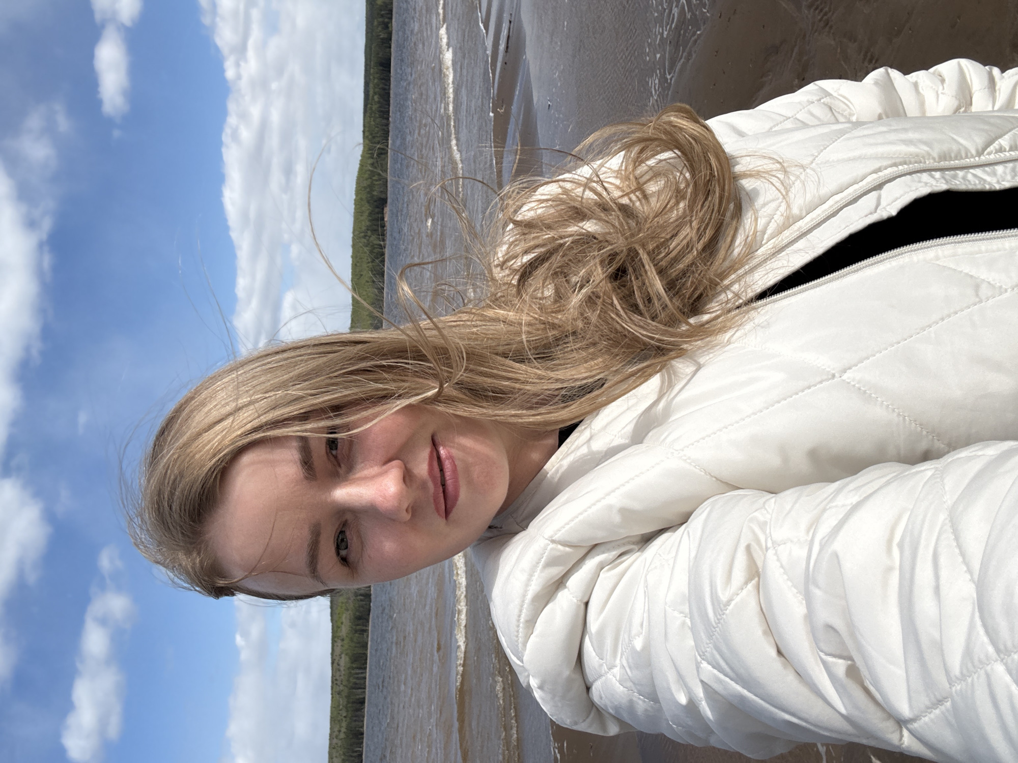 A young woman with blonde hair wearing a white puffy jacket is taking a selfie at the Kama Reservoir beach in Chaikovsky, Russia. The scene includes a cloudy blue sky, a shoreline with water waves, and a background with dense green trees on the opposite shore of the reservoir.