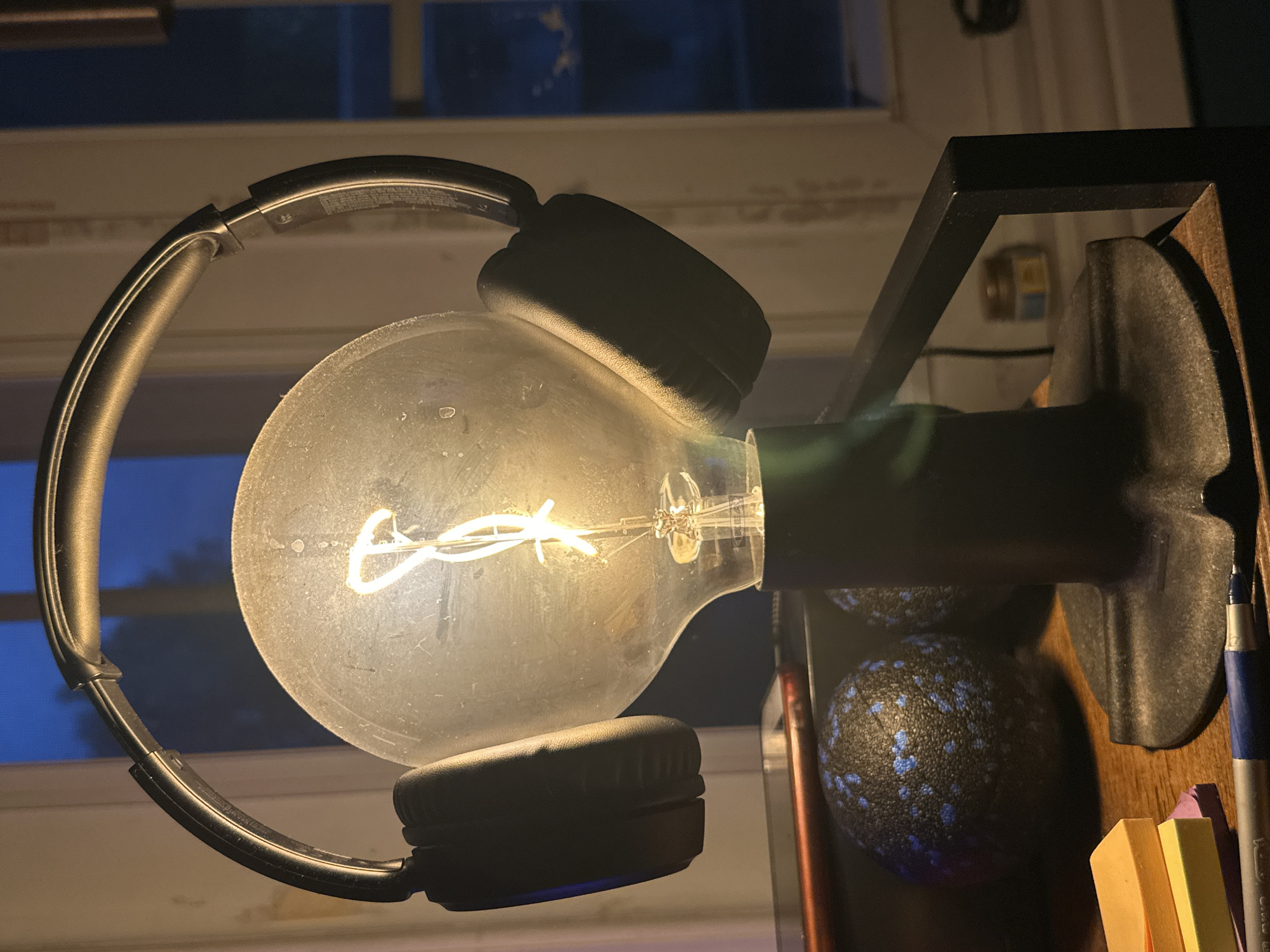 A close-up, low-angle shot captures a unique desk lamp in a dimly lit room, possibly a bedroom or office in McMasterville, Canada. The lamp features a clear, Edison-style light bulb with a visible filament, illuminated by a warm, yellow glow. The bulb is topped with a pair of black, over-ear headphones, the earcups resting on either side of the bulb. The headphones appear to be a modern, stylish accessory. The lamp itself has a dark, cylindrical base and a metallic stem, contributing to its industrial-chic aesthetic. In the background, out-of-focus, a window shows a deep blue twilight sky and a hint of a tree. On the desk in front of the lamp, there are several objects: a stack of colorful sticky notes (yellow, orange, and pink), a blue and silver pen, and two dark, textured spherical objects, possibly foam rollers. A dark, rectangular object, likely a phone or tablet, is partially visible behind the lamp. The overall impression is one of a personal space, perhaps a study area or a creative nook, decorated with eclectic items. The lighting creates a moody and intimate atmosphere.
