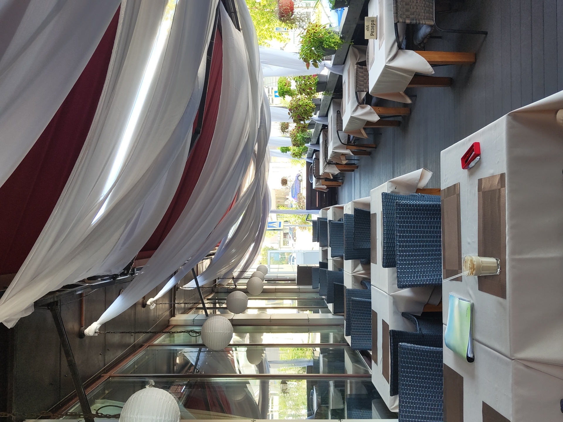 The image depicts the outdoor seating area of a restaurant in Zhukovsky, Russia, characterized by a covered patio with draped white and red fabric overhead. Several round, white paper lanterns hang from the ceiling. The patio is furnished with multiple tables, each covered with a light-colored tablecloth. The tables are set with placemats and chairs made of dark wicker. Some tables have reserved signs. On one table in the foreground, a light-colored beverage in a glass with a straw and a folder are visible. The scene suggests a well-maintained outdoor dining space.