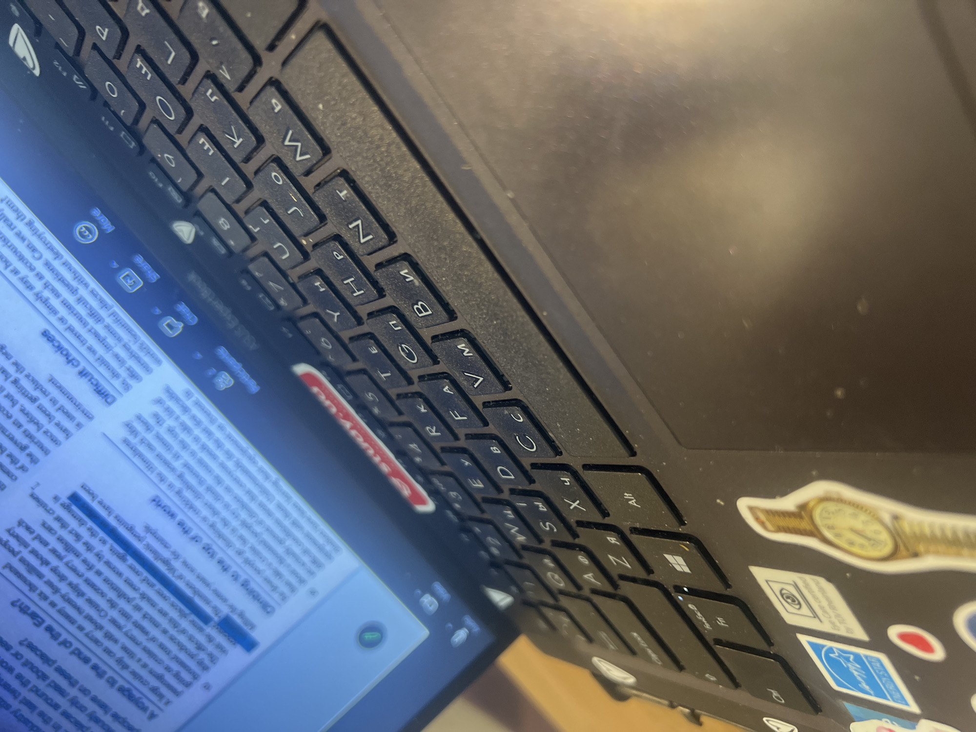 The image shows a close-up view of a laptop keyboard and a portion of the screen. The screen displays text, likely an article or book, with highlighted sections and the title "A voyage to the end of the Earth?". The keyboard is black and shows Cyrillic characters on some keys, indicating a non-English keyboard layout. There are various stickers on the laptop casing, including a watch sticker, an "ENERGY STAR" logo, and others. The scene appears to be indoors, with artificial lighting. There are no people visible in the frame. The overall impression is one of someone working or studying on a laptop. The location is not discernible from the image itself.