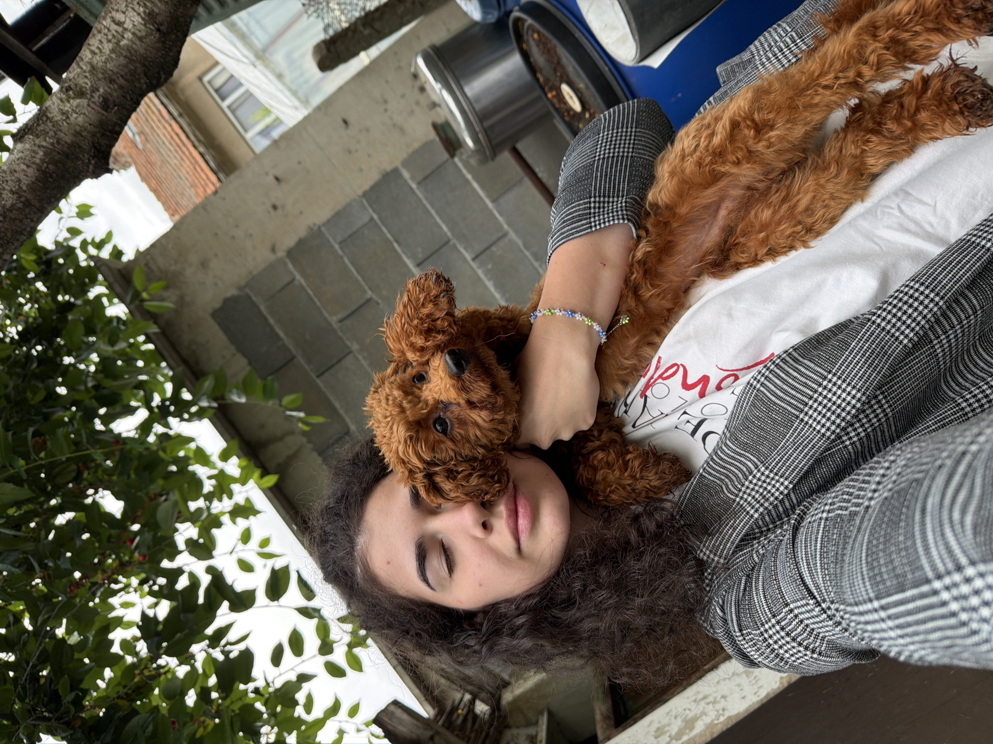 In Sofia, Bulgaria, a young woman with dark, curly hair and closed eyes is taking a selfie outdoors, holding a small, reddish-brown poodle. She is wearing a grey and black plaid jacket over a white t-shirt with red lettering. The dog, with its ears perked, is held close to her face. Behind them, a concrete block wall and a tree with green leaves are visible. Further in the background, a red-tiled roof of a building and a greenhouse suggest a residential or rural setting. Several large plastic containers and a metal object are also present in the background, possibly indicating a yard or storage area.