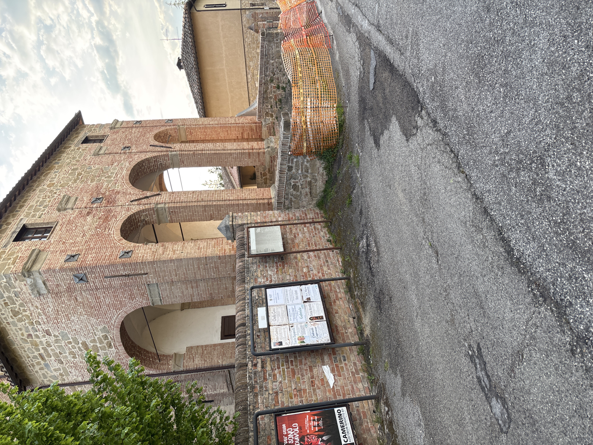 The image, captured in Camerino, Italy, shows a historic street scene dominated by a multi-story building constructed from brick and stone. The building features several arched openings, some with views into interior spaces or passages, and smaller rectangular windows. Details like metal X-shaped wall anchors and varied brickwork patterns are visible on its facade. Green foliage from a tree is present in the lower left foreground.
To the right, a paved road with visible cracks and minor repairs curves gently. A bright orange plastic construction or safety fence runs along the edge of the road, separating it from low stone walls and parts of the building structure.
Multiple information boards are mounted on the brick wall. One poster at the bottom advertises "GREG GREGORI SONO DIAVOLO" and mentions "CAMERINO AUDITORIUM BENEDETTO XIII," indicating a local event. Above it, a larger board displays several individual notices, likely obituaries or public announcements, with names such as Pietro Canfrivi, Orello Cioci, Lucio Tapanelli, Gabriele, Teresa Martella, and Gabriele Capocasa discernable. Another smaller, seemingly blank board is higher up.
The scene takes place during the daytime under an overcast sky, providing soft, diffused lighting. No people are visible, suggesting a quiet moment in this historic town setting. The image is presented rotated 90 degrees counter-clockwise from its original capture orientation.