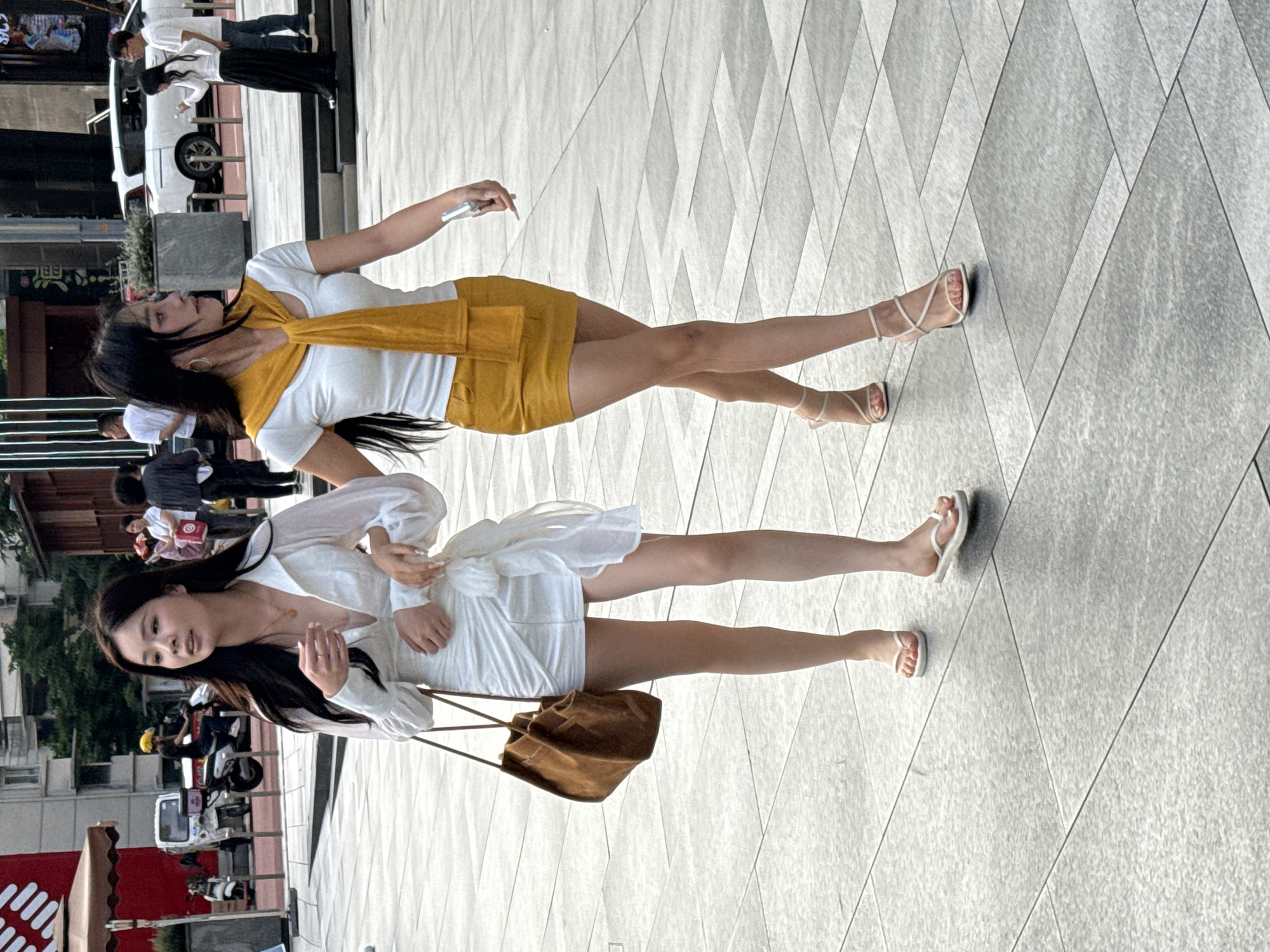 Two young women are standing on a tiled urban walkway, posing for a photograph. One woman wears a white short-sleeved top with a yellow scarf, and a yellow skirt, and sandals. The second woman wears a white dress with long sleeves, carries a brown shoulder bag, and wears flip-flops. The location appears to be in a public pedestrian area within the city of Liangjiang, China. People, vehicles, and buildings are visible in the blurred background, indicating an active urban environment.