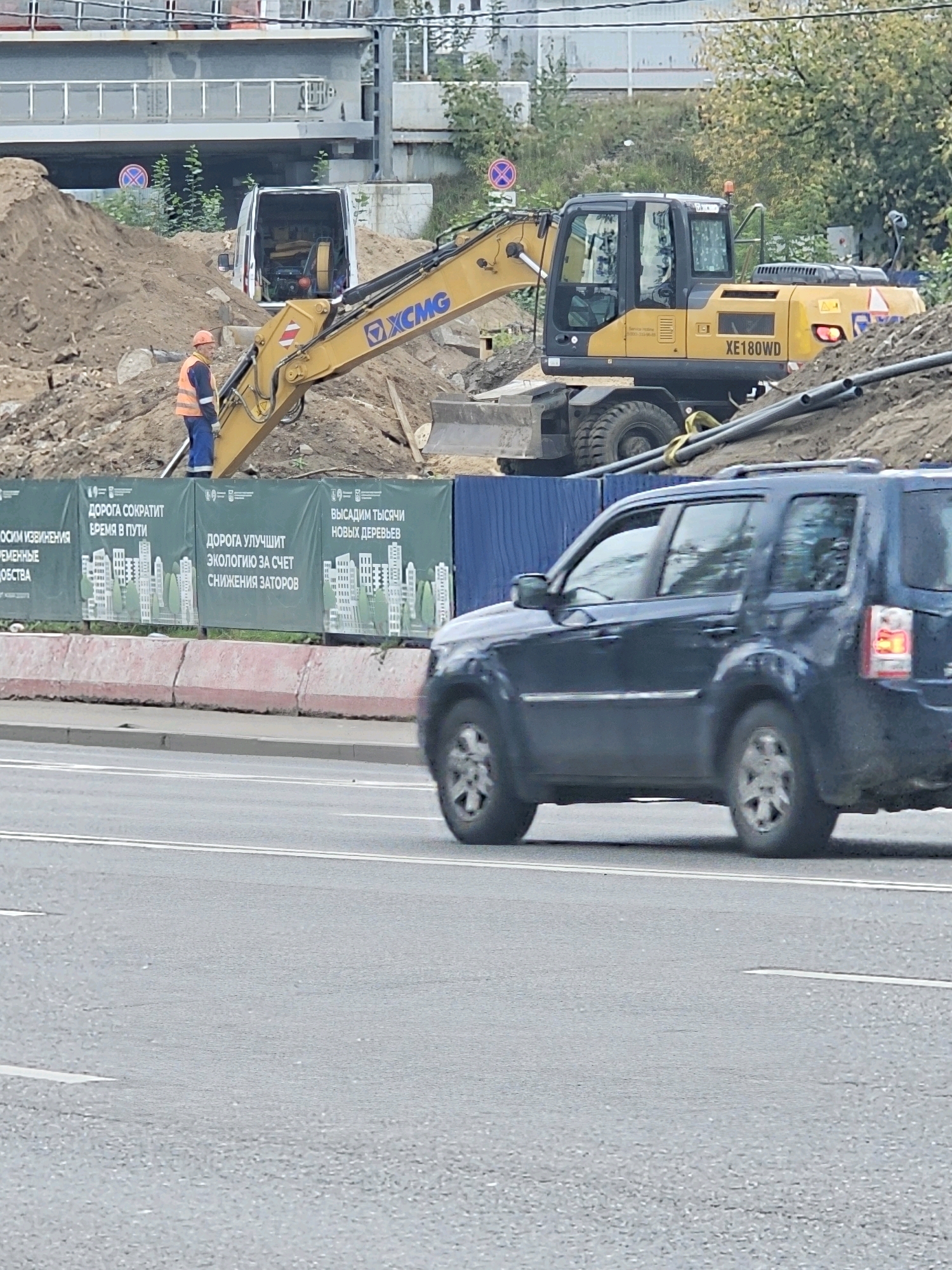 The image shows a construction site with a yellow XCMG excavator in the background. A construction worker in an orange vest and blue pants stands near the excavator. A white van is parked behind a pile of dirt. A dark blue SUV is driving on a road in the foreground. A green banner with text and images of buildings is stretched across the construction site. The scene appears to be a road construction project in Moscow, Russia, with a focus on improving infrastructure and the environment.