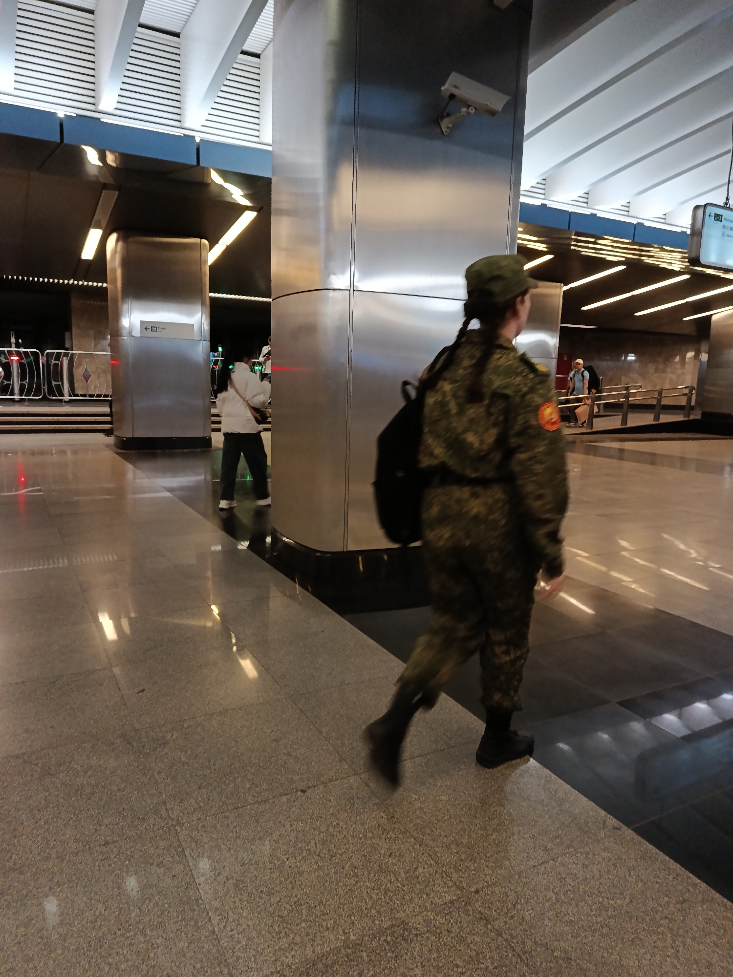 A person in a military uniform and cap walks through a subway station. The person is wearing a camouflage uniform and black boots, with a backpack slung over their shoulder. Their hair is in a braid. The station has polished tile flooring and metal support beams, with a surveillance camera mounted on one of the beams. Other people are visible in the background. The location is most likely in the city of Moscow, Russia, based on the appearance of the station and the uniform worn by the person.