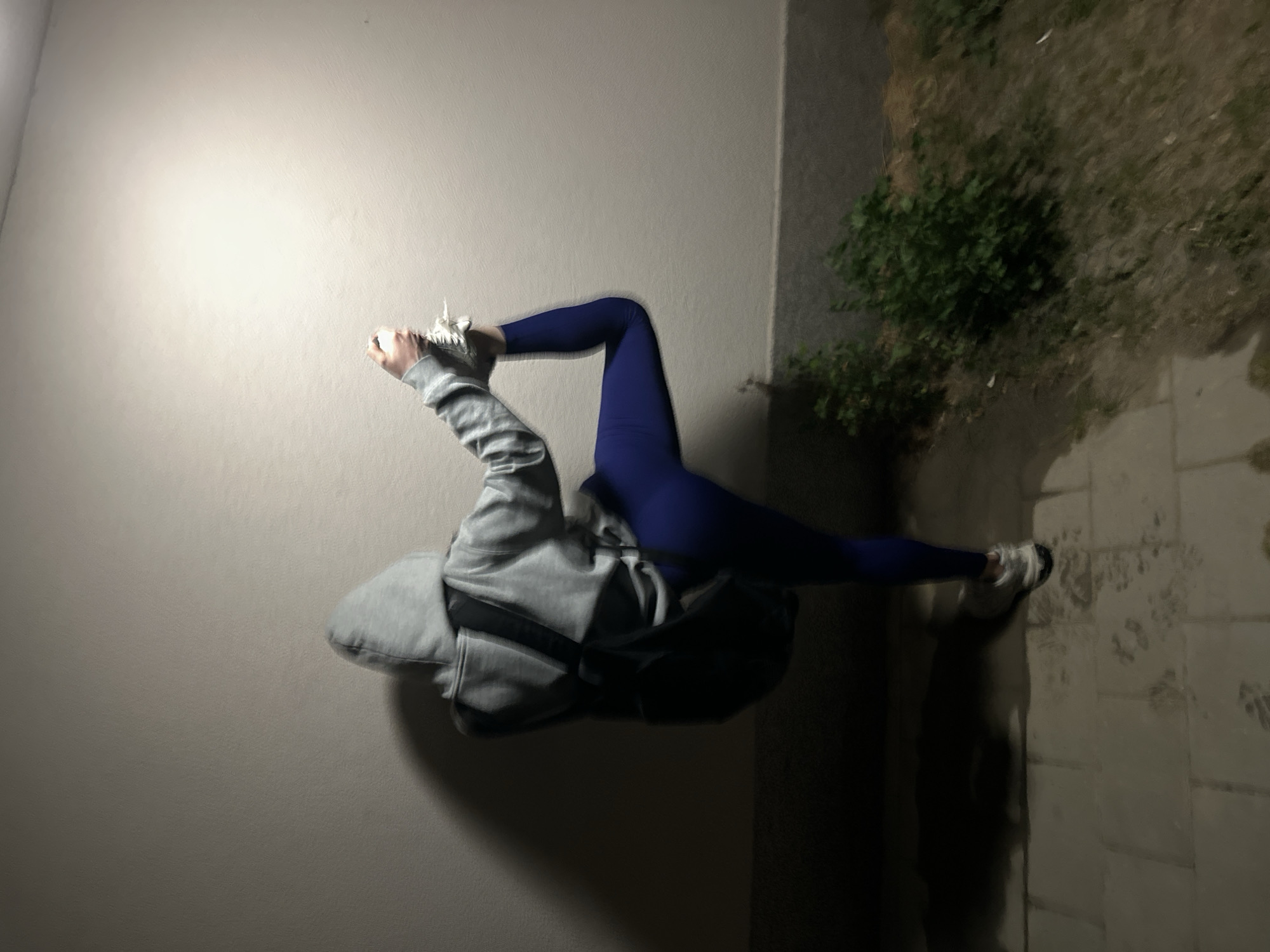 A person in a gray hooded sweatshirt and blue leggings is performing a contortionist pose outdoors. They are positioned with their back on a light-colored wall, and one leg is extended towards a sidewalk made of light-colored pavers. The other leg is bent at the knee and raised towards the wall. The person is wearing white sneakers on both feet. The sidewalk is adjacent to a patch of greenery with some light visible in the upper right. The location is potentially in Sofia, Bulgaria, due to the query's focus, but this is not verifiable from the image alone. The person is the main subject, and the action is a physical pose. The environment consists of a wall, sidewalk, and greenery. Interactions are limited to the person's pose and interaction with their surroundings.