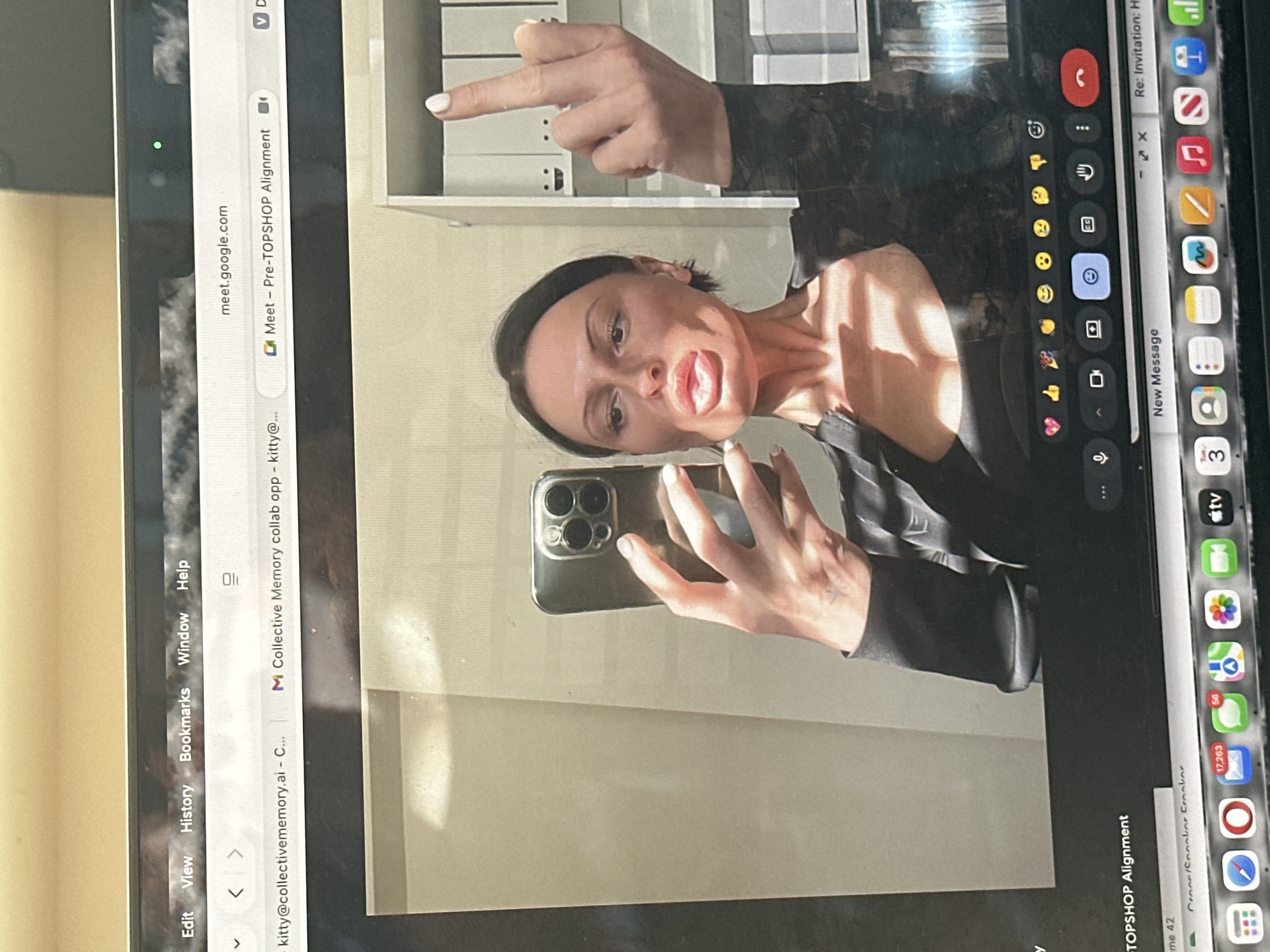 The image is a photograph of a laptop screen displaying a Google Meet video call. The screen's upper section shows browser tabs, including "meet.google.com" and a tab titled "Meet - Pre-TOPSHOP Alignment." The primary subject of the video call is a woman with dark hair pulled back, wearing a dark v-neck top. She holds a dark multi-lens smartphone in her left hand, appearing to record or take a selfie, while puckering her lips. Her right hand is raised with the middle finger extended. The background behind her features a light-colored wall and a white shelving unit containing several white binders, one of which has a simple black smiley face drawn on its spine. Below the video frame, the Google Meet interface shows various control icons and reaction emojis. The laptop's macOS dock is visible at the bottom of the screen, displaying app icons such as Messages with 17,263 unread notifications and WhatsApp with 56 unread notifications. The specific physical location within Wealden, United Kingdom, cannot be determined from the visual information presented in the image.