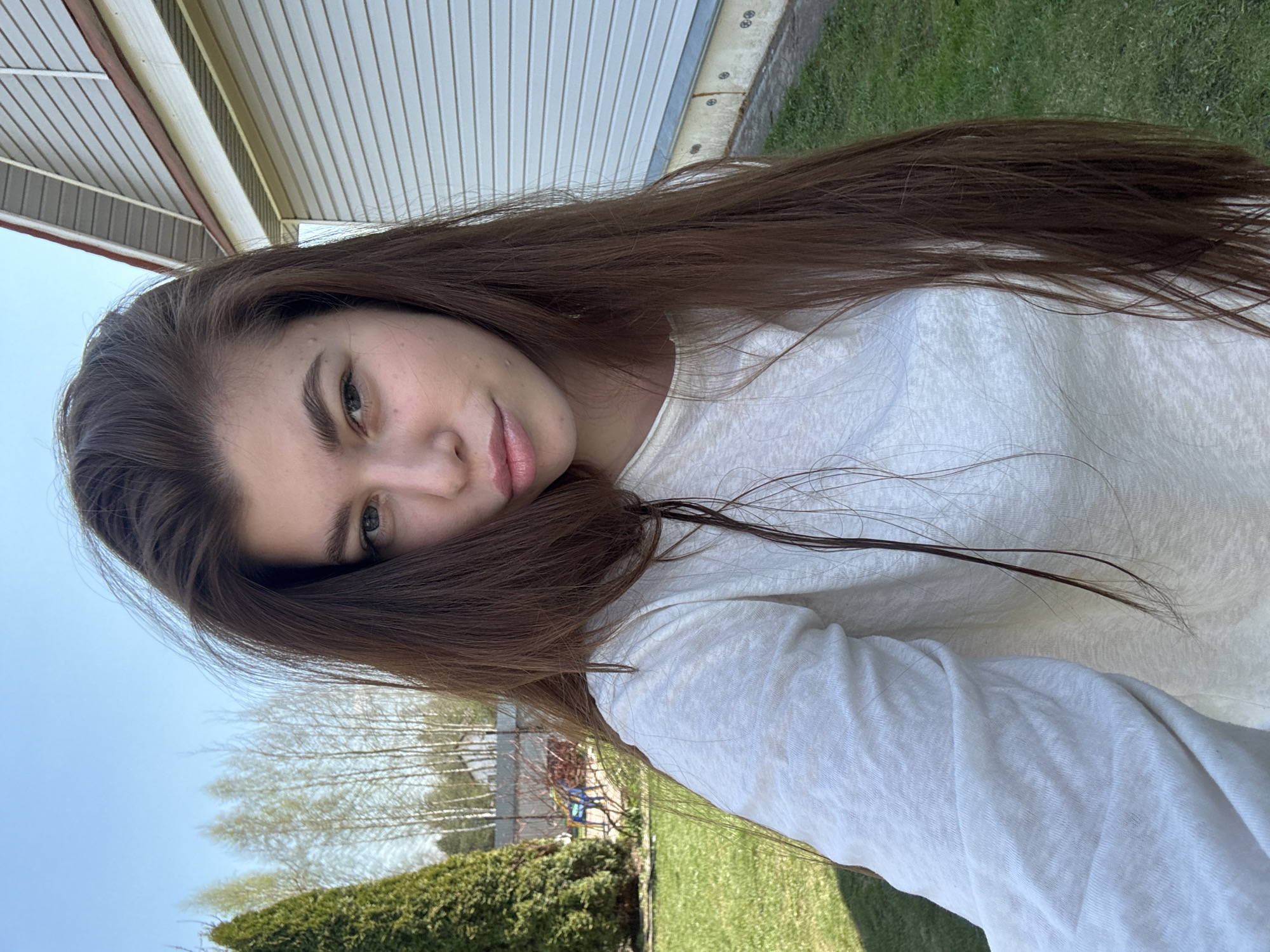 A young woman with long brown hair is the subject of a selfie taken outdoors. Her face is partially obscured by her hair. She is wearing a white, long-sleeved shirt. Behind her, the background includes a house with white siding, green grass, trees, and a blue sky. No information is available in the image to confirm the location as Мещёры, Russia.