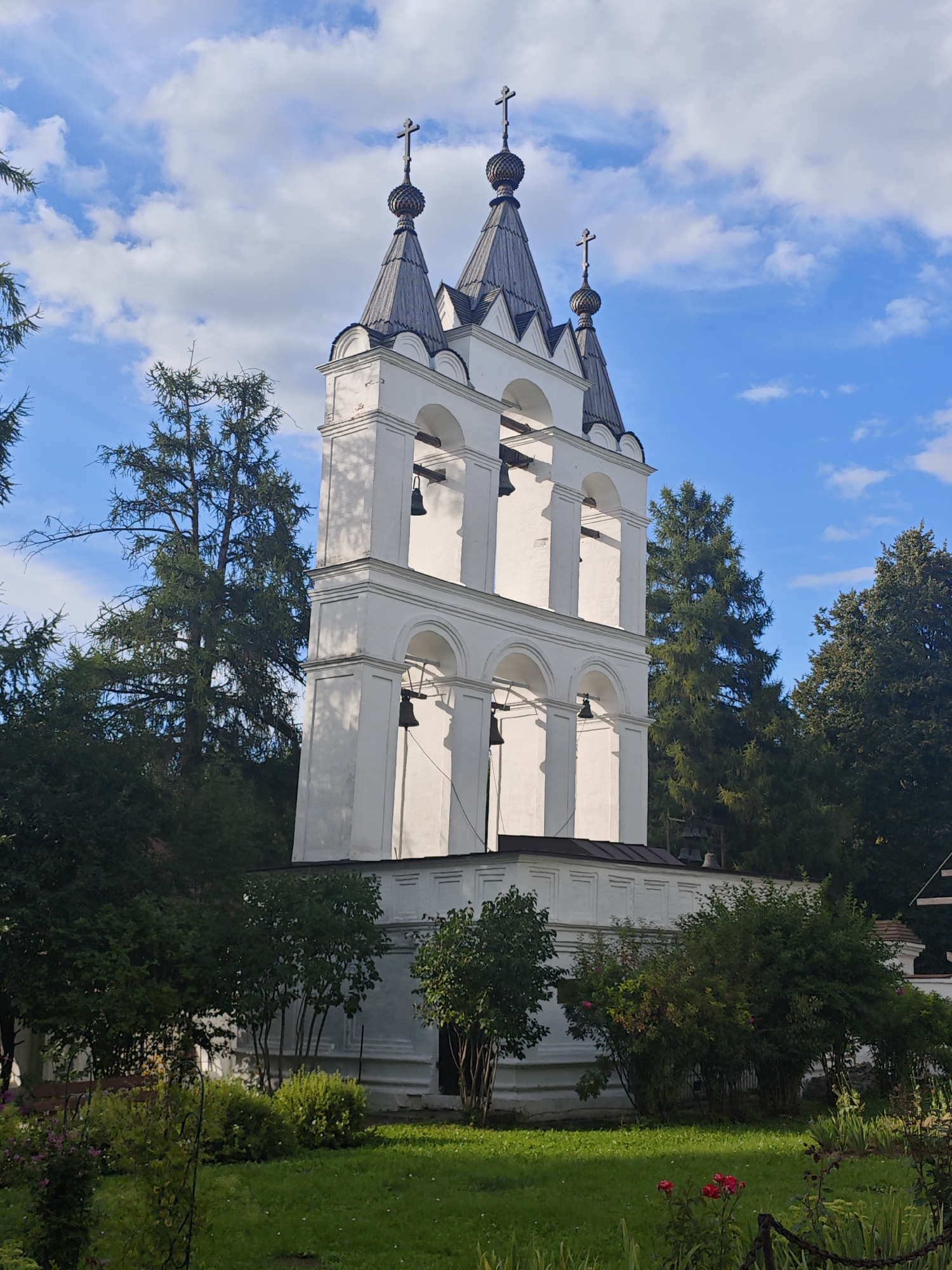 A white bell tower with three tiers and a dark, multi-pitched roof with multiple spires topped with crosses stands against a blue sky with scattered white clouds. The bell tower is surrounded by lush green trees and foliage. The lower portion of the bell tower has arched openings with bells hanging inside. The scene is illuminated by sunlight, casting shadows on the tower and the surrounding greenery. In the foreground, there is a neatly trimmed lawn with some flowering plants. The location is identified as Bolshiye Vyazyomy, Russia, suggesting this is likely a religious building within a historical or monastic complex. There are no people or animals visible in the image.
