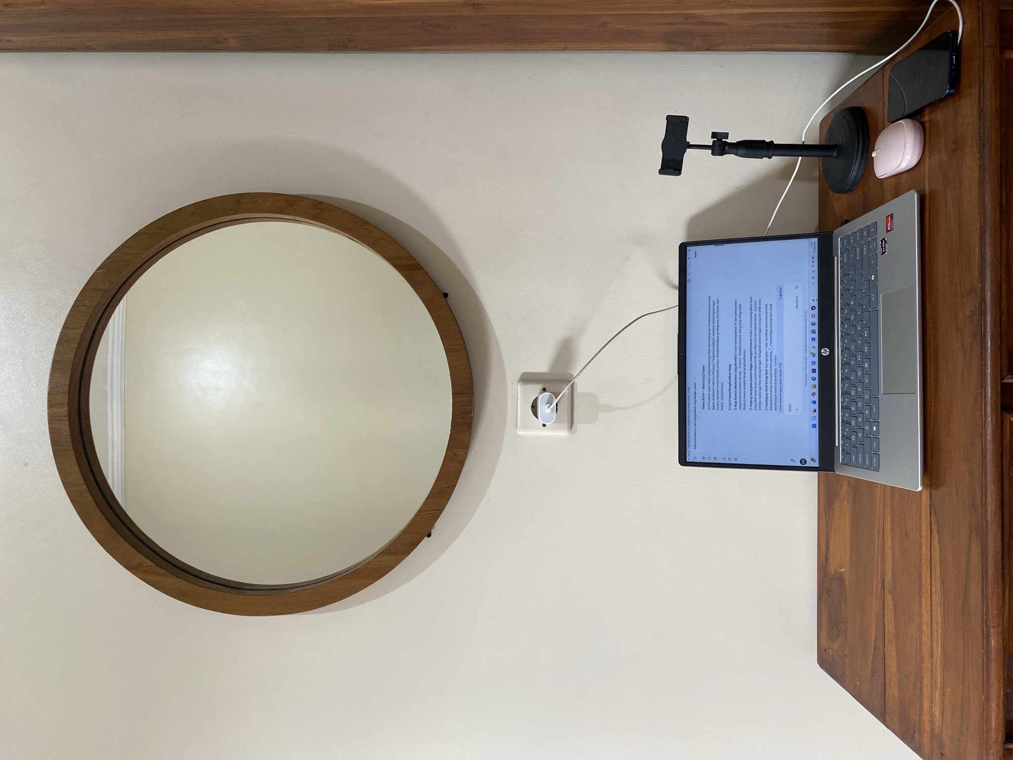 The image depicts a desk setup against a light beige wall. A round wooden-framed mirror hangs on the wall to the left. Adjacent to the mirror, a white power outlet is visible with a charging cable plugged in. To the right of the mirror and outlet, there is a laptop open on the desk, displaying a document. Further right, there is a phone stand, a light pink computer mouse, and a smartphone charging on the desk's right edge. The desk has a rich brown, varnished wood finish. The items and the background suggest an indoor workspace.
Based on the document on the laptop screen appearing to be written in Indonesian, this image was most likely taken in Indonesia. Specifically, the first line on the laptop screen references "Mata Uang Utama," which translates to "Main Currency," and this may indicate that the text is from a news article discussing cryptocurrency. Based on the image content, it is not possible to identify the exact location in North Kuta.