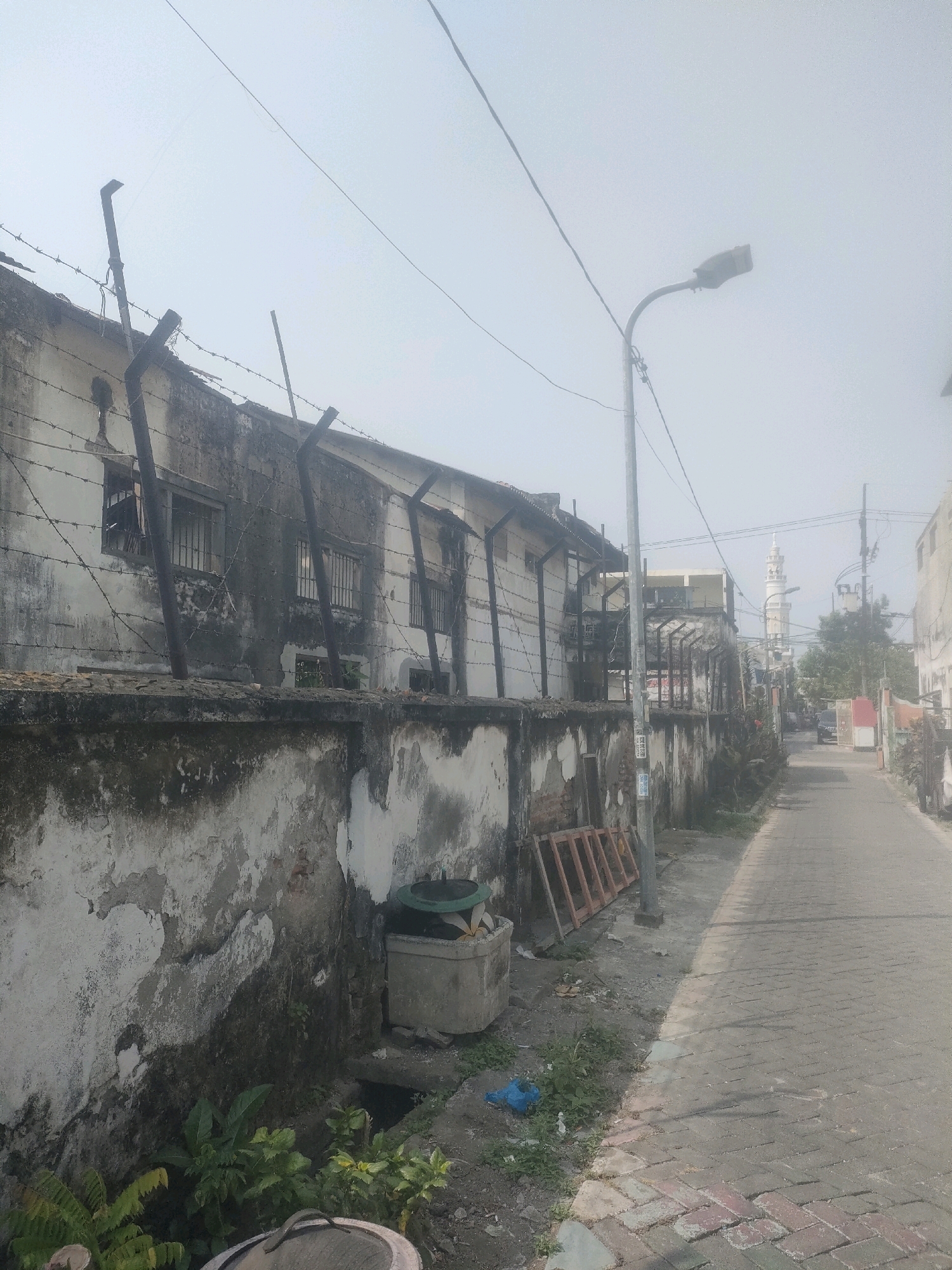 The image shows a narrow, paved street in Surabaya, Indonesia, bordered by a dilapidated concrete wall and a multi-story building with barred windows. The building appears to be an older structure, with peeling paint and visible signs of disrepair. Barbed wire runs along the top of the wall and the building's roofline, suggesting a security concern or a former restricted area. A street lamp stands on the right side of the road, its light obscured by the hazy atmosphere. Further down the street, a mosque minaret is visible in the distance, indicating a predominantly Muslim neighborhood. The overall scene suggests an urban environment characterized by aging infrastructure and a somewhat neglected appearance. The sky is overcast, and the air appears hazy, possibly due to pollution or fog. There are no people visible in the image.