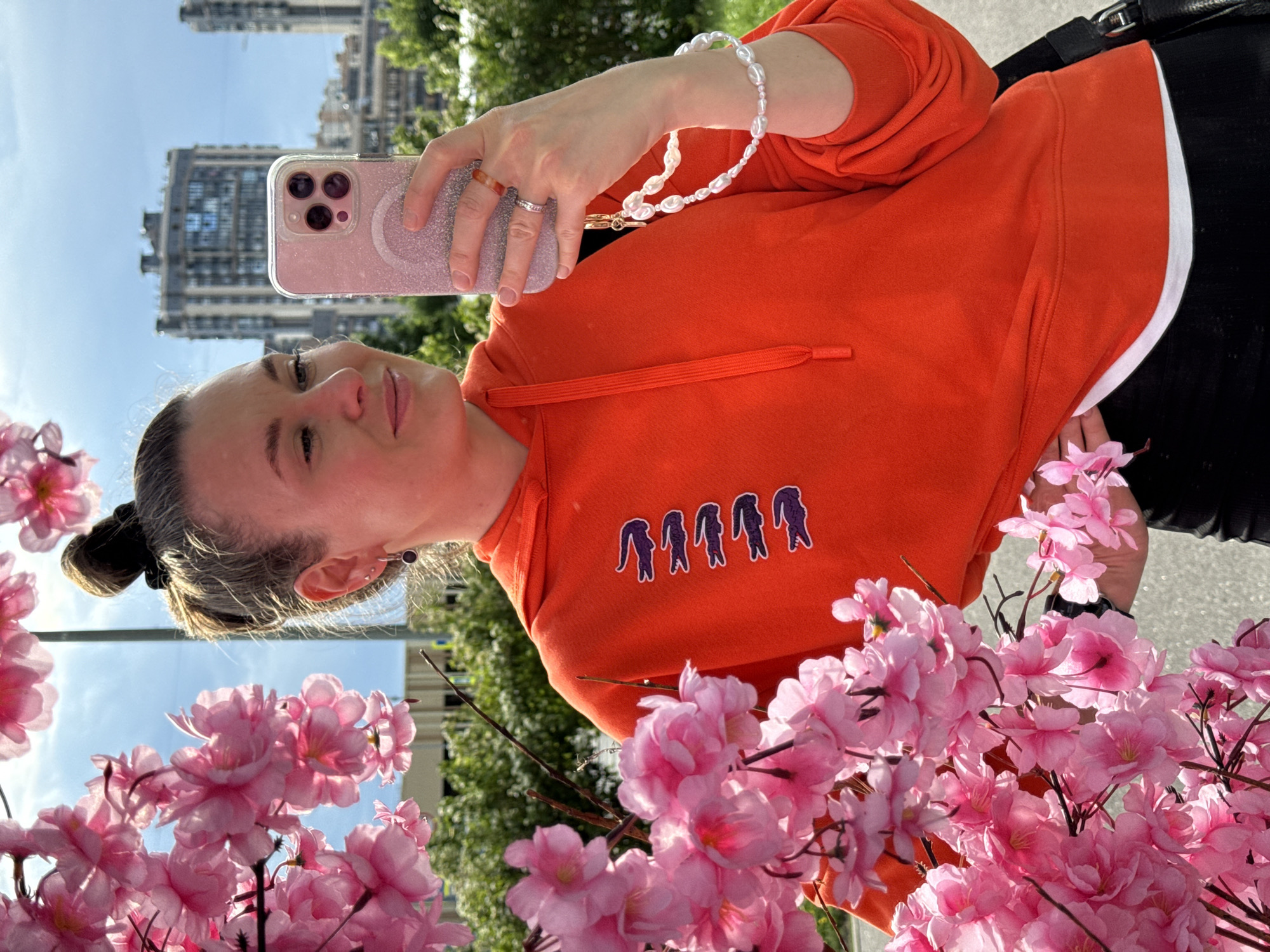 A woman is taking a selfie with a smartphone while holding a branch of pink artificial cherry blossoms in front of her. She is wearing an orange hoodie with a string of purple alligator logos down the left side, and a white t-shirt underneath. She has a pearl bracelet on her left wrist and two rings on her right hand. The background shows a bright blue sky with some clouds, a row of modern apartment buildings, and green trees. The woman's hair is pulled back into a bun, and she has subtle makeup on. The photo appears to have been taken outdoors in an urban environment, possibly in Saint Petersburg, Russia, given the architectural style of the buildings.
