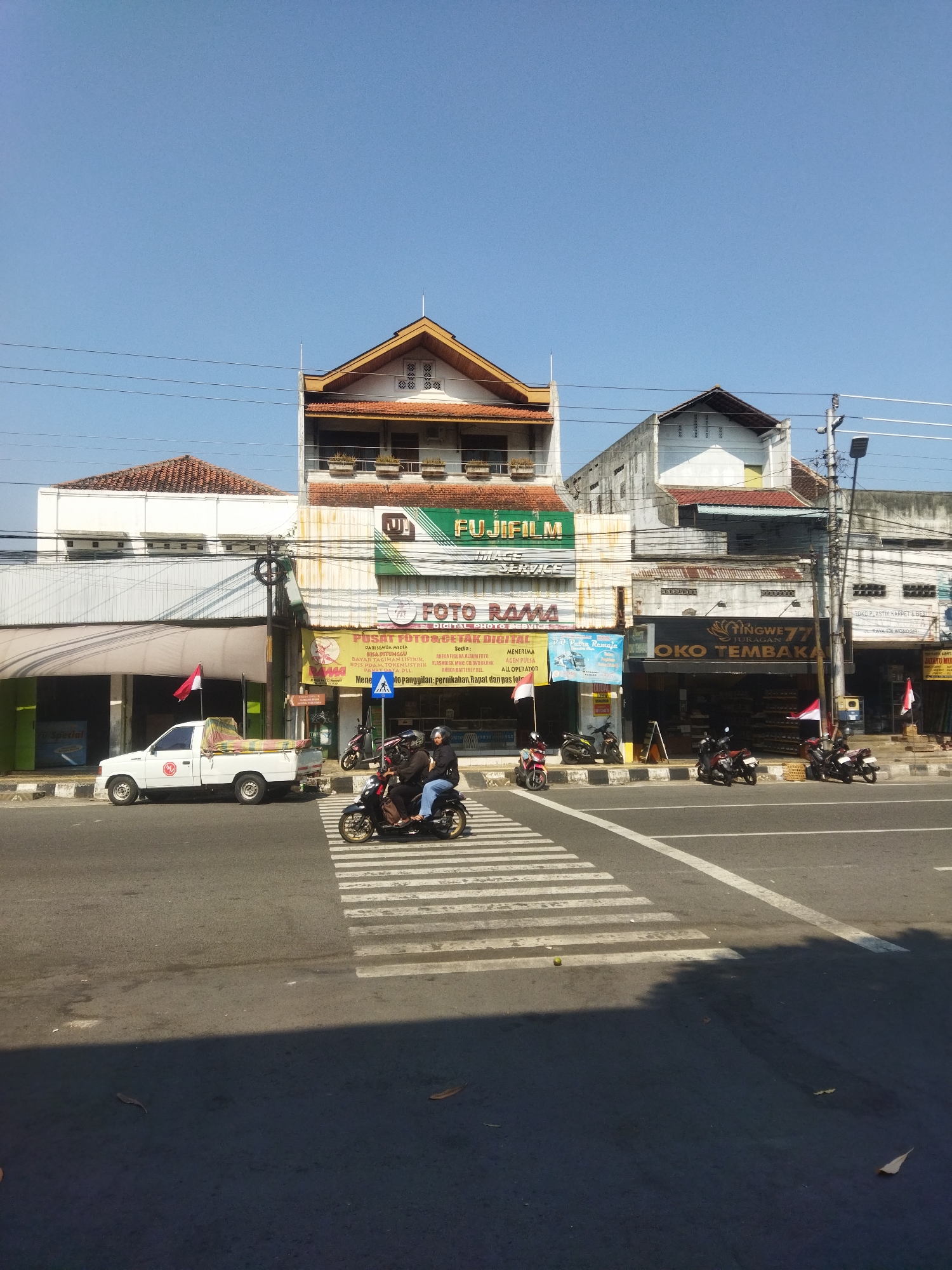 This is a photograph taken in the city of Wonogiri, Indonesia, on a clear, sunny day. The scene depicts a street with a pedestrian crossing in the foreground. A white pickup truck is parked on the left side of the road, and two people are riding a motorcycle on the pedestrian crossing. Several other motorcycles are parked along the sidewalk in front of shops. The buildings lining the street are commercial establishments, including "FUJIFILM", "FOTO RAMA", and "OKO TEMBAKA". Indonesian flags are displayed in front of some of the shops. The sky is a bright, clear blue, and the sun is casting shadows on the road. The overall impression is that of a typical day in a bustling Indonesian town.