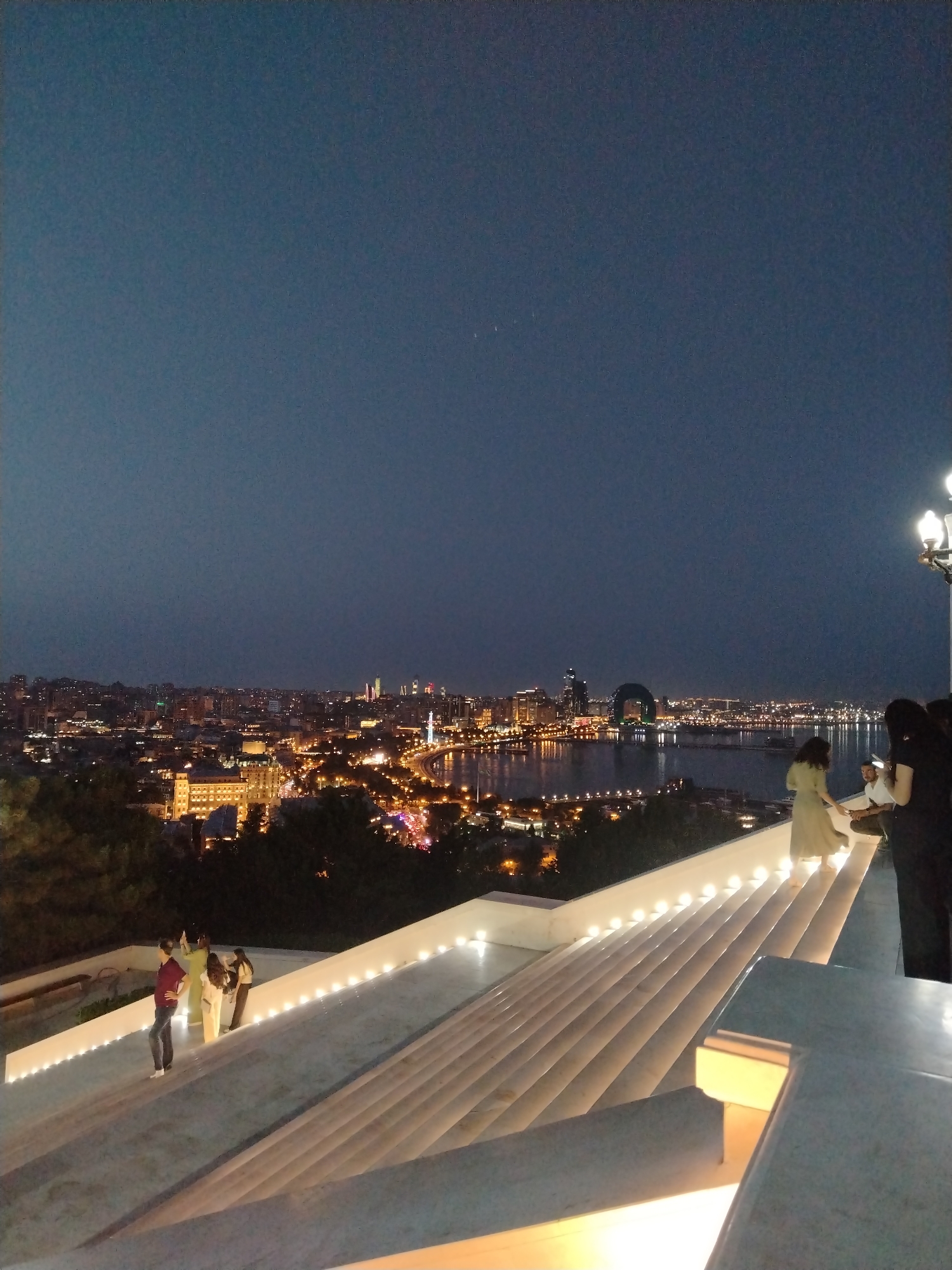 The image shows a panoramic view of the city of Baku at night from a viewpoint in Sabail Raion, Azerbaijan. In the foreground, a set of wide, modern white stairs with integrated lighting leads down from the viewpoint. Several people are visible on the stairs, some standing and looking at the city, others in conversation. A man in a maroon shirt and jeans stands on the left side of the stairs, facing away from the camera. Further up the stairs, a woman in a light-colored dress and two other individuals are standing. In the background, the Baku cityscape stretches out, illuminated by numerous lights. The Caspian Sea is visible in the middle ground, reflecting the city lights. Prominent landmarks include the Flame Towers and the curved building with a loop, also known as the Flame Towers or Baku City). The sky above is dark blue, indicative of twilight or night. The overall scene depicts a popular tourist or viewing area offering a scenic overlook of the city.