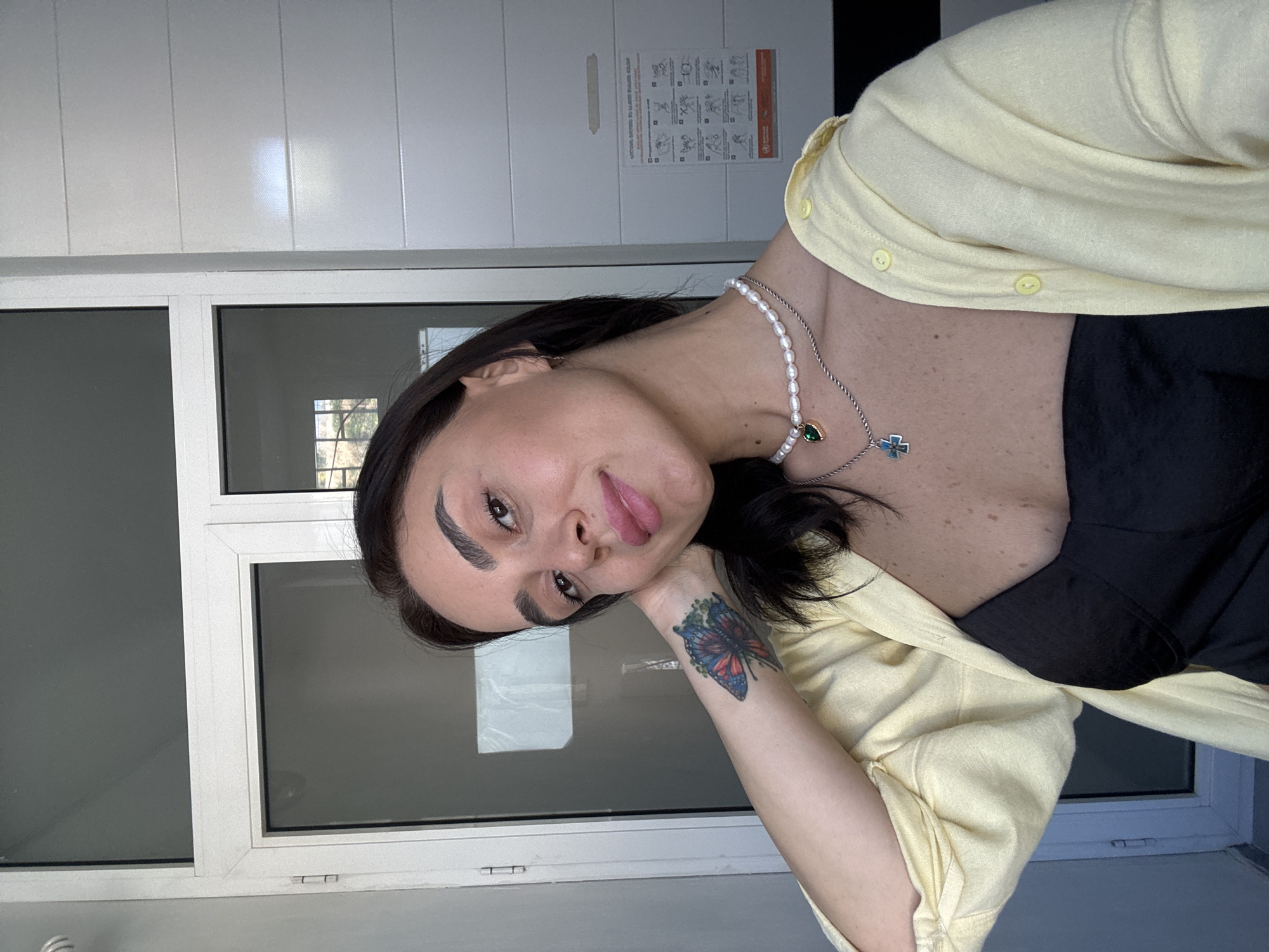 A young woman with dark hair and fair skin poses for a selfie in what appears to be an indoor setting with large windows and a textured wall. She is wearing a black strapless top and a pale yellow cardigan. She has a butterfly tattoo on her left forearm and is wearing two necklaces, one with a pearl strand and a green pendant, and another with a silver chain and a blue cross pendant. Her left hand is resting on the back of her head. The background consists of a large window or door with white framing, and a wall with horizontal textured panels. There is also a white safety poster with diagrams on the textured wall. The overall lighting suggests it is daytime. The scene does not contain specific elements that definitively place it in Tbilisi, Georgia.