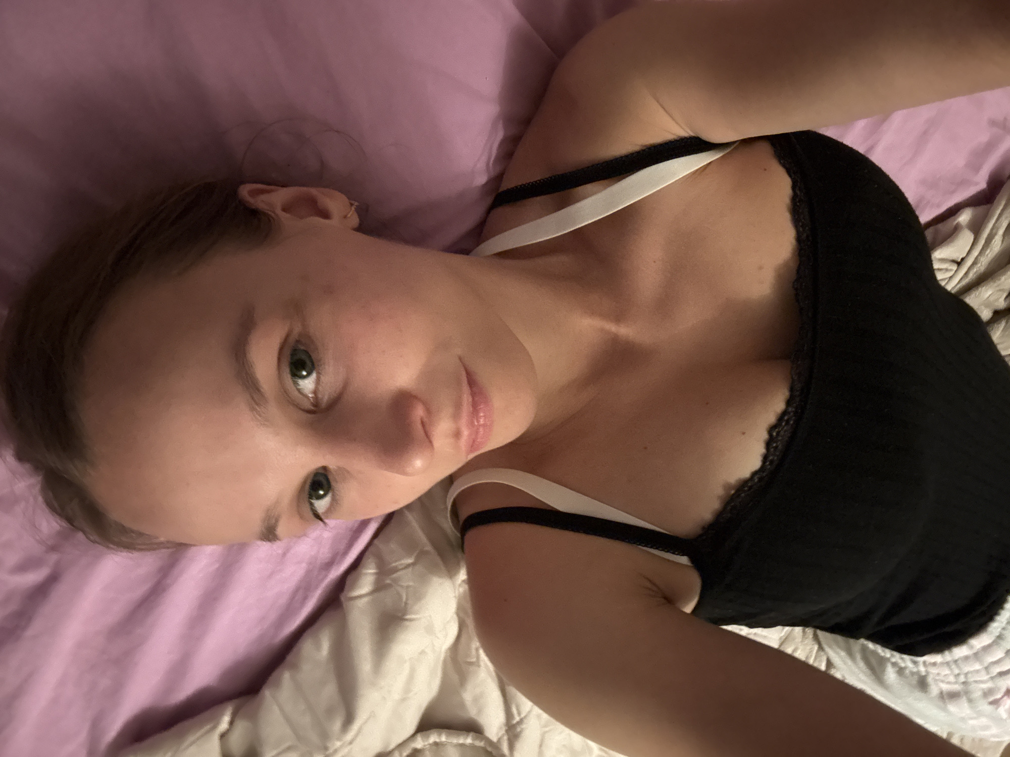 A woman is lying on a bed with purple sheets. She is wearing a black tank top with white straps and white shorts. She has light brown hair and green eyes. The woman is looking at the camera and smiling. The room is dimly lit, with the exception of the light coming from the camera flash. The bed appears to be in a bedroom. There is no visible indication of the location being in Überherrn, Germany.