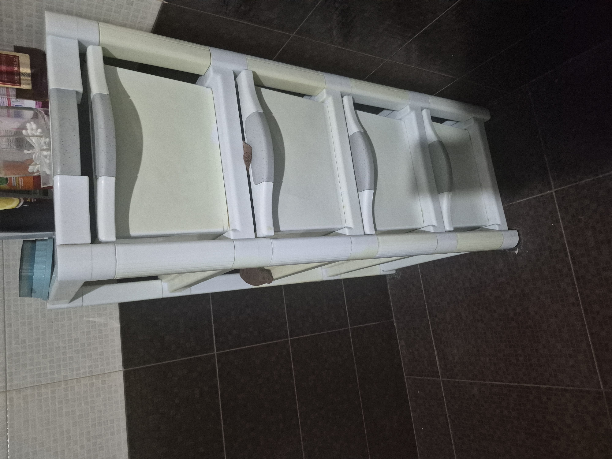 The media file displays a white, multi-tiered plastic storage unit, likely a chest of drawers, situated in an indoor room with tiled surfaces. The unit comprises five compartments, four of which are fully visible. Each compartment features a light off-white or cream-colored front panel and a distinctive gray, ergonomic handle that appears textured and shows signs of use or dirt.
To the immediate left of the storage unit, on an adjacent shelf or cabinet, various items are visible. These include a bottle with a golden label, bearing partially visible text like "100%", and several clear plastic containers holding small objects, possibly bathroom accessories or personal care items. A blue translucent container is also partially visible at the bottom left.
The setting suggests a functional space, such as a bathroom, laundry room, or utility area, located in Yerevan, Armenia. The floor is covered with dark brown or black square tiles, while the wall behind the unit features lighter, possibly beige or white, small mosaic-patterned tiles. The scene is static, illuminated by artificial indoor lighting, and no people or specific activities are depicted.