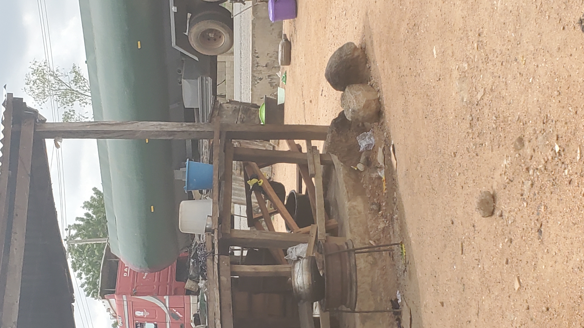 The image depicts an outdoor scene in Jalingo, Nigeria. A large, green cylindrical tank, likely part of a tanker truck, is prominently featured, supported by a wooden structure. The truck's red cab is partially visible below the tank. Various objects are scattered around the area, including buckets, rocks, and what appears to be trash. A wooden scaffold or shelter is also present, with some corrugated metal roofing. The ground is sandy and uneven, with small stones and debris. The sky is overcast, suggesting a cloudy day. The overall impression is one of a somewhat cluttered, utilitarian space, possibly a truck stop or a service area. There is no visible text in the image.