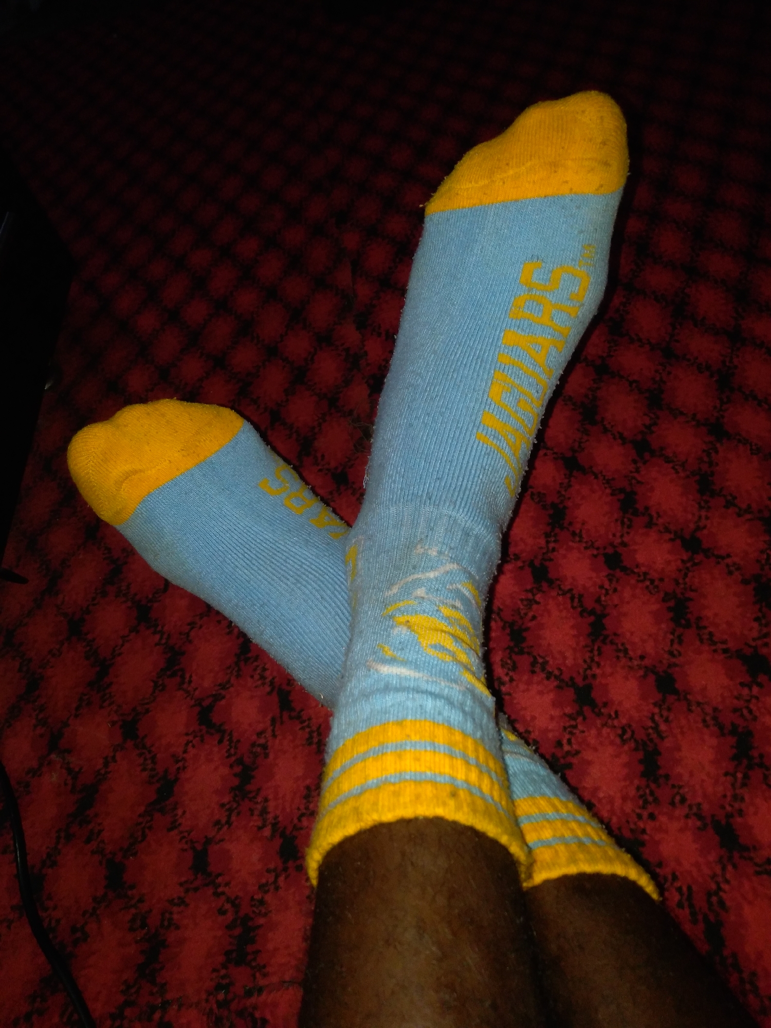 The image shows two light blue socks with yellow toe caps, heels, and ribbed cuffs. The word "JIMARS" is visible in yellow lettering on one of the socks. The socks are worn by a person whose legs are visible. The person is lying down on a red and black checkered surface, possibly a blanket or carpet. The scene is indoors and dimly lit.