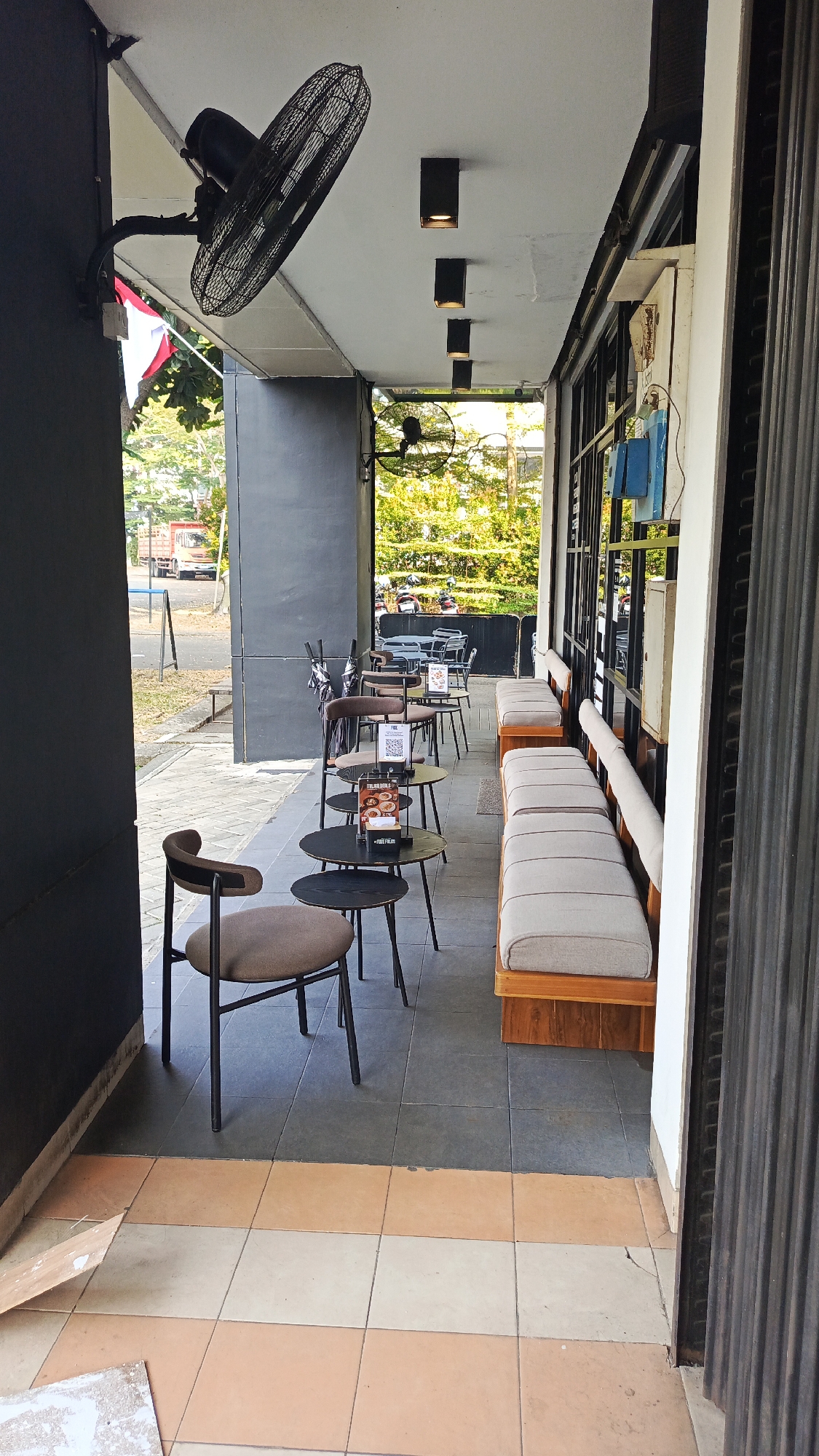 The image shows an outdoor seating area of a cafe or restaurant. The area is covered by a roof, and there are several tables and chairs set up for customers. In the foreground, there is a brown chair with a black metal frame, and to its right, a series of small, round black tables are arranged. Some of these tables have menus or other items on them. Further back, more tables and chairs are visible, including some umbrella stands. Along the right side of the seating area, there is a long, built-in bench with light-colored cushions, which appears to be part of the building's structure. Above the seating area, a large black industrial fan is mounted to the wall. The ceiling has several rectangular light fixtures.
The background reveals a street scene. To the left, a road is visible with a red truck in the distance. Trees and foliage can be seen beyond the seating area. The overall impression is that of an urban environment, likely in a commercial district, given the cafe-style seating and street access. Based on the context provided, this scene is located in Tangerang, Indonesia.