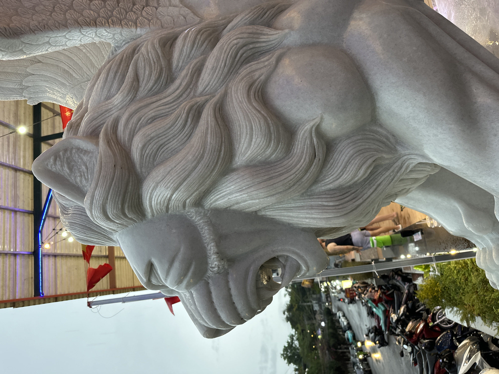 A white marble statue of a winged lion dominates the frame. The statue has a detailed mane and an open mouth, revealing its teeth. In the background, an outdoor setting shows a street scene with parked motorcycles lining the side of the road. Green trees and foliage are visible along the street. A person wearing a green and grey workout outfit is also visible, partially obscured by the statue. Red flags and overhead string lights are visible in the distance. Based on the provided location of Bac Nha Trang Ward, Vietnam, it suggests that the statue is located in this area.