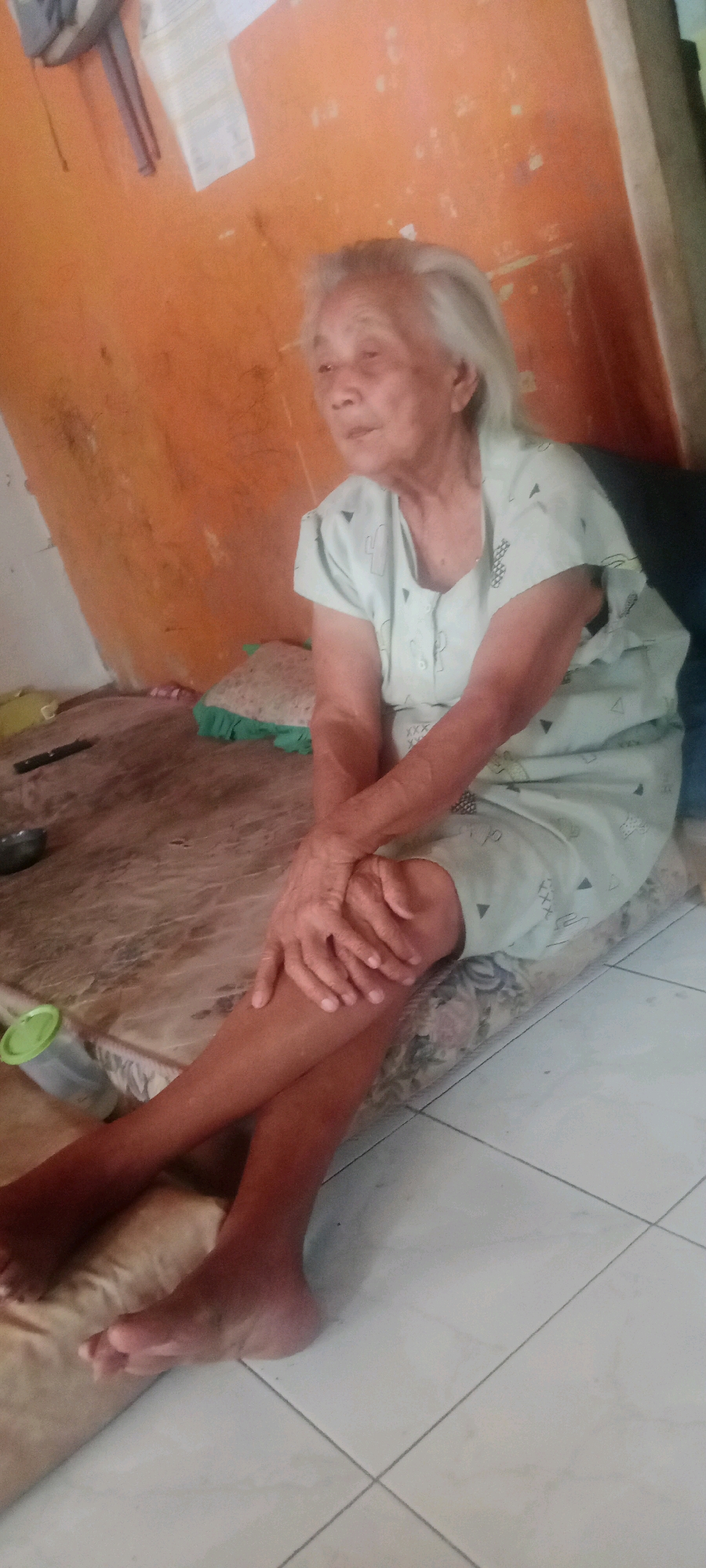 An elderly woman with light-colored hair sits on a mattress on the floor, facing right with her legs crossed. She is wearing a light green, short-sleeved dress with a subtle cactus pattern and white buttons down the front. Her hands are clasped over her left knee. The mattress is placed on a wooden floor. Behind her, an orange-colored wall is visible, with some papers or fabric hanging on the upper left. To the right of the woman, a dark-colored object, possibly a couch or chair, is partially visible. The floor in front of the mattress is tiled in a light color. A green and white plastic cup is on the left side of the mattress. The scene appears to be indoors, likely in a residential setting in the city of Kasihan, Indonesia.