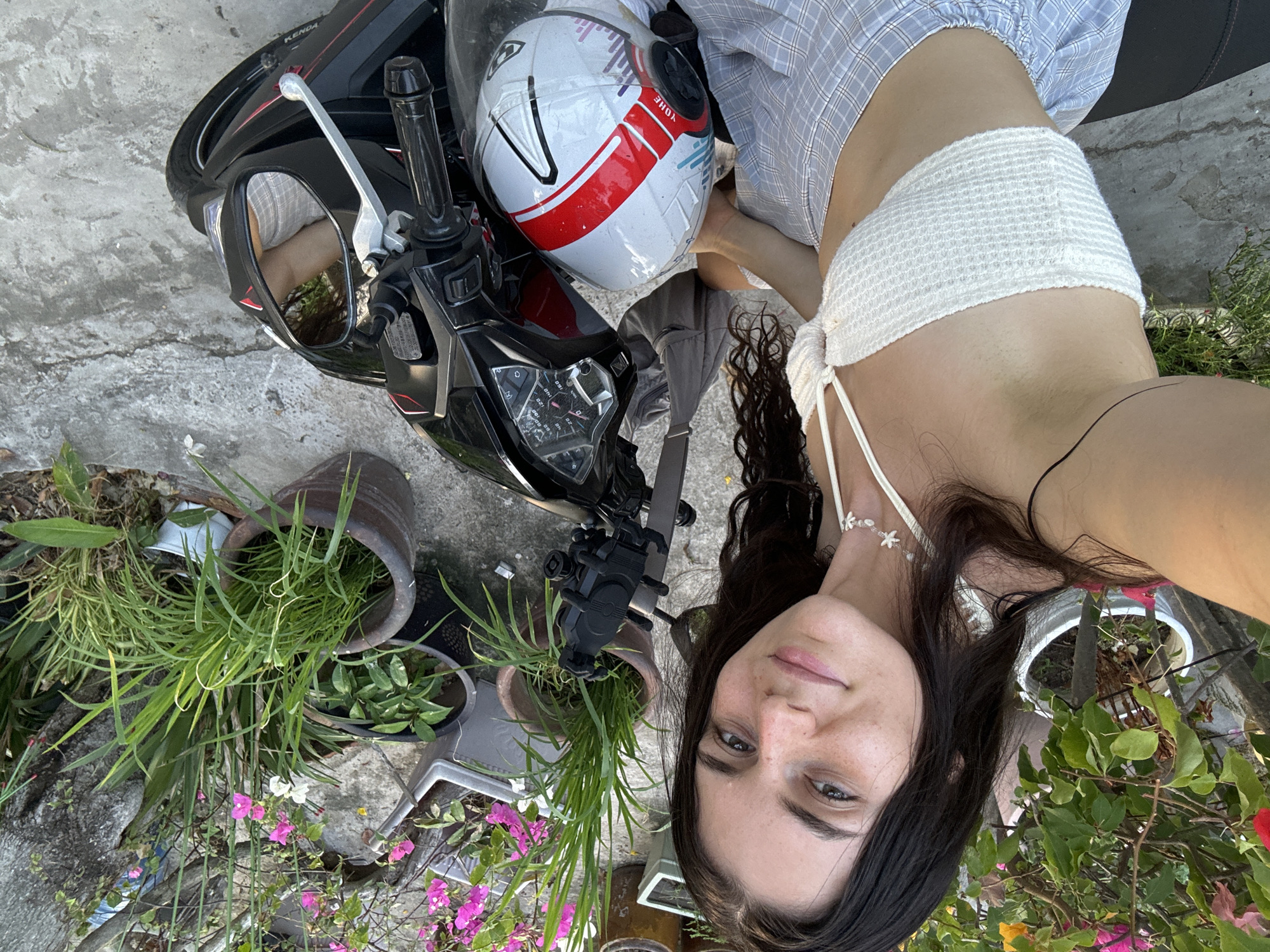 A selfie depicts a woman adjacent to a motorcycle in an outdoor setting with plants and pots. The motorcycle, predominantly black with a white and red helmet resting on it, is adjacent to several potted plants with green foliage and pink flowers. The woman, with dark hair and fair skin, is wearing a white crocheted top and a light blue patterned shirt. Her gaze is directed towards the camera. The image suggests a setting in Mũi Né, Vietnam based on the vegetation and general landscape.
