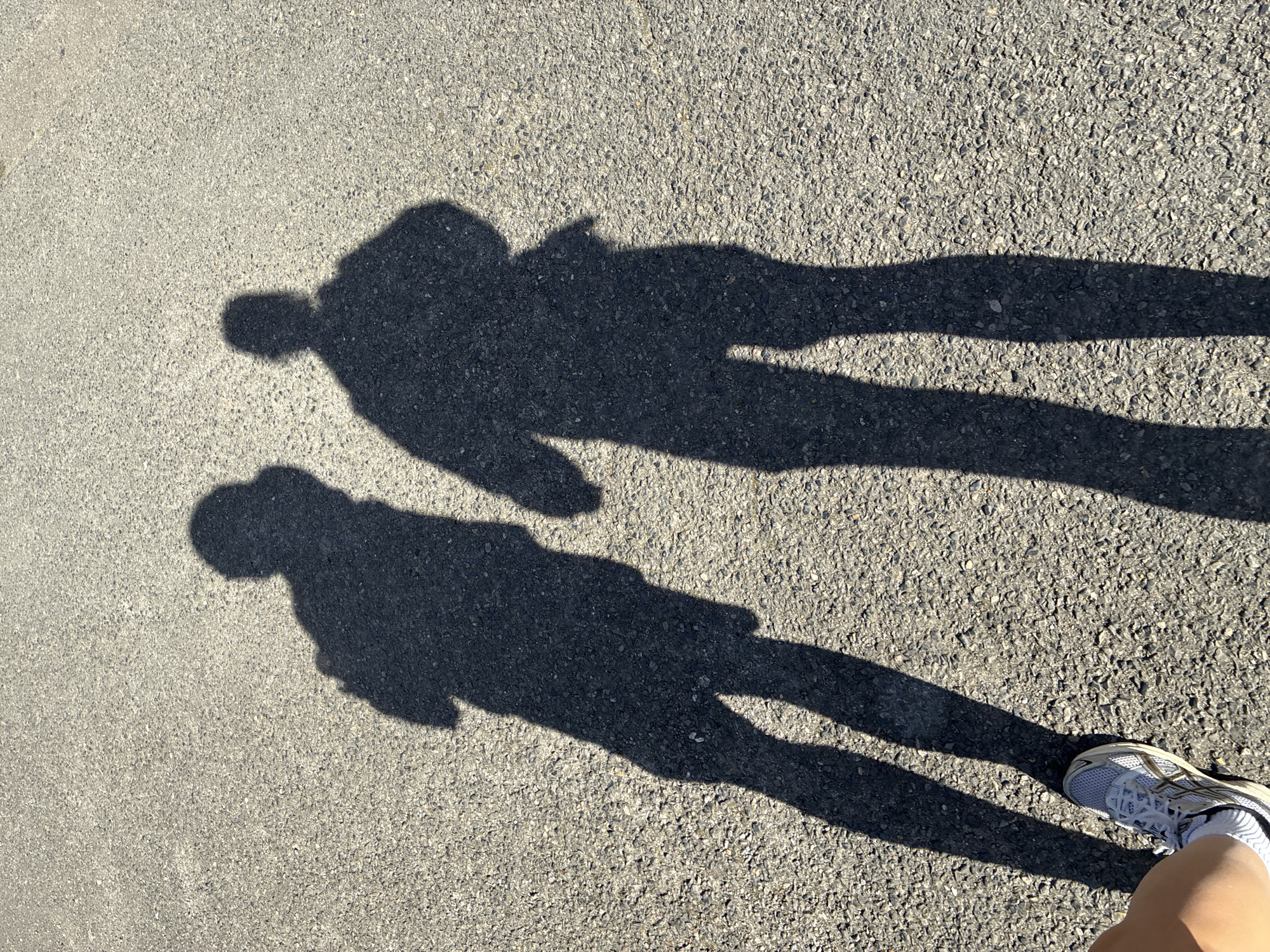 The image depicts two shadows on an asphalt surface, likely a road or pavement. One shadow appears to be of a person wearing a backpack, while the other shadow seems to be of a person in motion, possibly walking or running. A portion of a leg and shoe are visible in the lower right corner, indicating the person taking the photograph. The environment is an outdoor urban setting, as suggested by the asphalt surface, and the presence of the shadows indicates it is daytime with sunlight. The image doesn't contain indicators of a particular location in London.