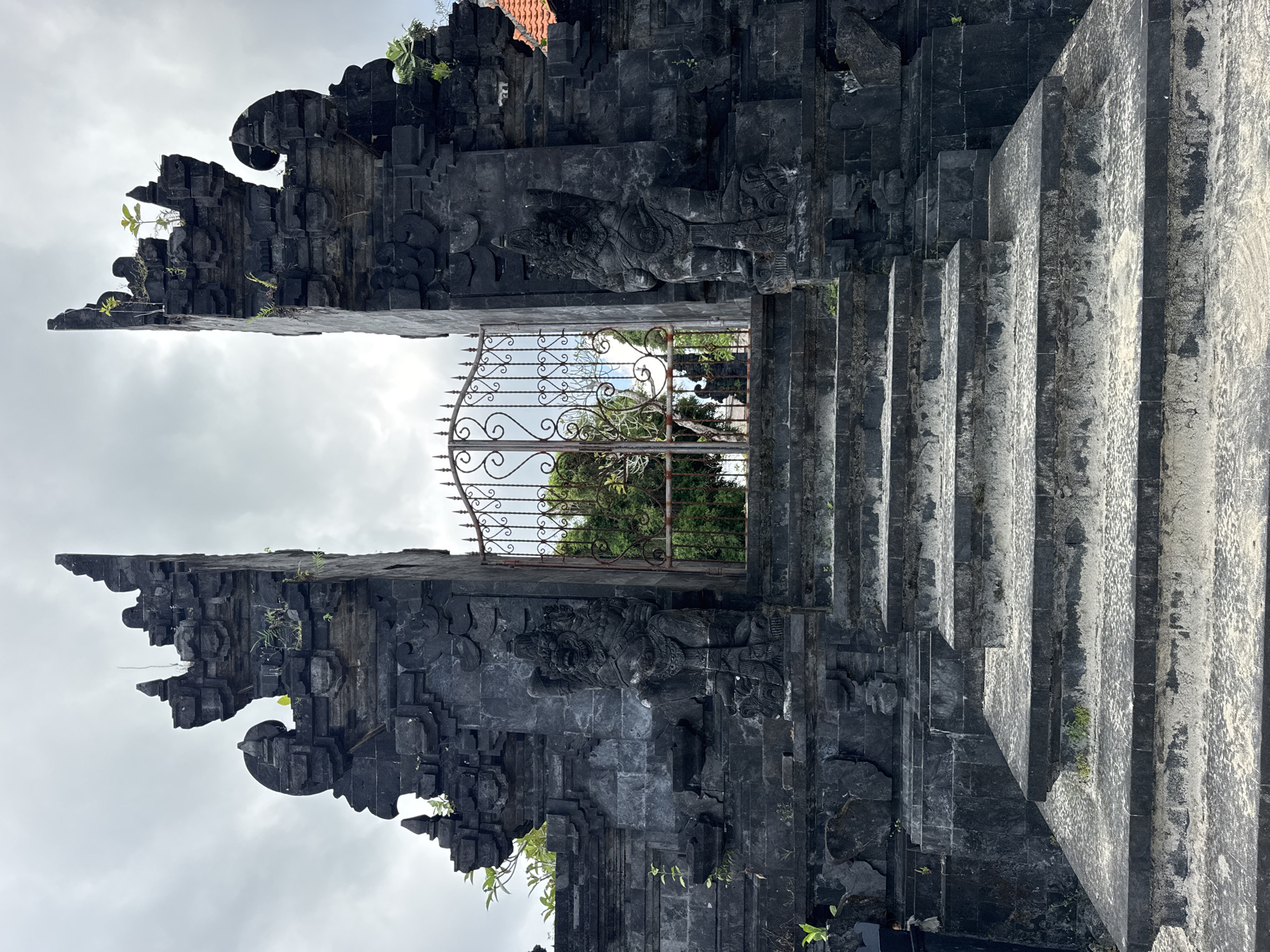 This image depicts the elaborate stone gate of a Balinese Hindu temple. The gate is constructed from dark-colored stone, possibly volcanic rock, with intricate carvings and ornate detailing. The architectural style features traditional Balinese elements, including tiered structures and sculptural ornamentation. A wrought-iron gate stands between the two pillars of the larger stone structure. The environment appears to be overgrown with greenery. This temple is located in Kuta Selatan, Indonesia.