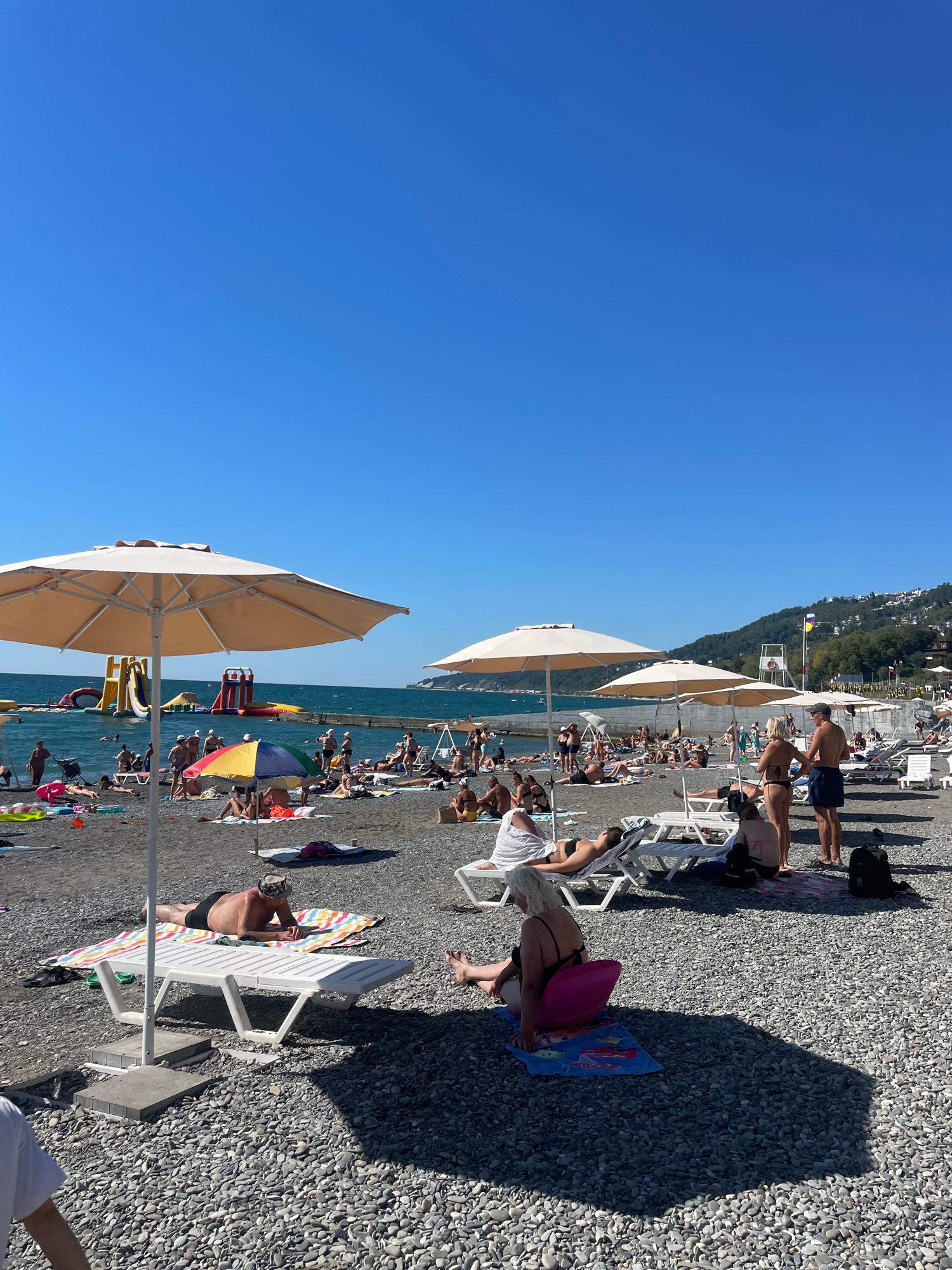 This image depicts a crowded pebble beach in Sochi, Russia, on a clear, sunny day. The foreground is dominated by a large, light-colored beach umbrella. Beyond that, the beach is populated with numerous sunbathers, many reclining on lounge chairs or towels. In the mid-ground, a colorful umbrella shields a group of people. The sea is visible in the background, with several inflatable water slides and structures in the water, indicating a recreational area. The coastline curves to the right, with buildings and vegetation visible on the hillside. The overall scene is one of leisure and activity on a typical summer day at the beach.