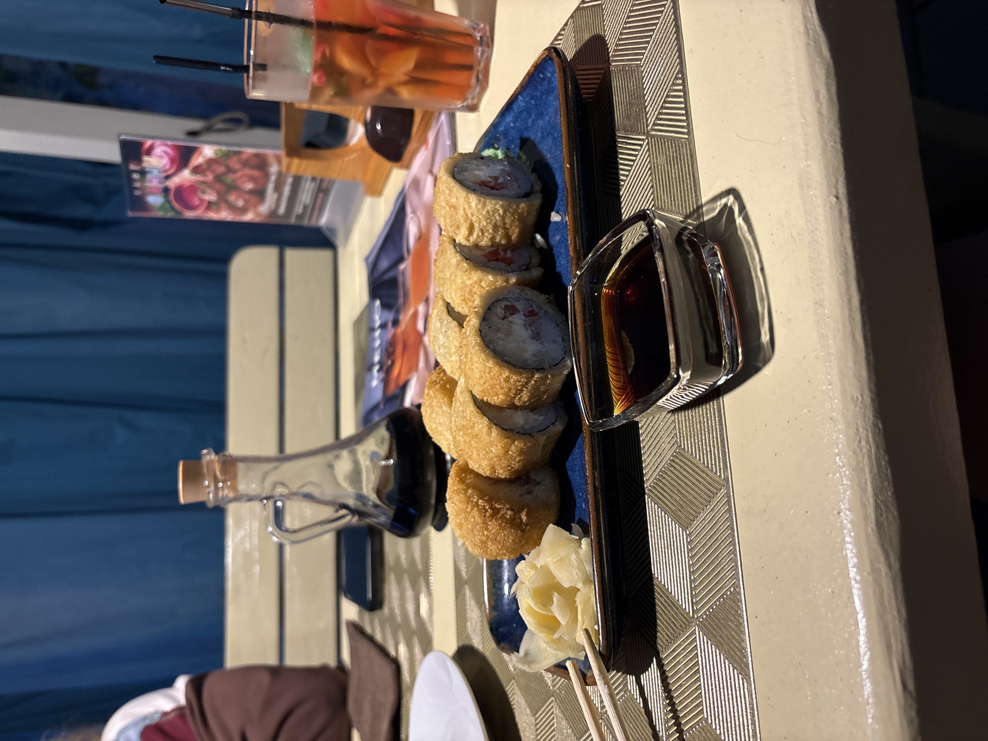 In the city of Chaikovsky, Russia, a table is set with a plate of tempura sushi rolls, pickled ginger, chopsticks, a small dish of soy sauce, and a glass of a fruity beverage. The sushi rolls are golden brown and appear to be filled with a creamy white substance and red ingredients. The pickled ginger is pale yellow and shredded. The soy sauce is dark brown and in a clear glass dish. The beverage in the glass is a reddish-orange color with ice, fruit pieces, and mint leaves, garnished with two black straws. A small glass carafe with a cork stopper, containing dark liquid, is positioned behind the sushi. In the background, blue curtains are visible. The table surface has a textured pattern. A portion of a person, wearing a brown garment, is visible on the left side of the image.