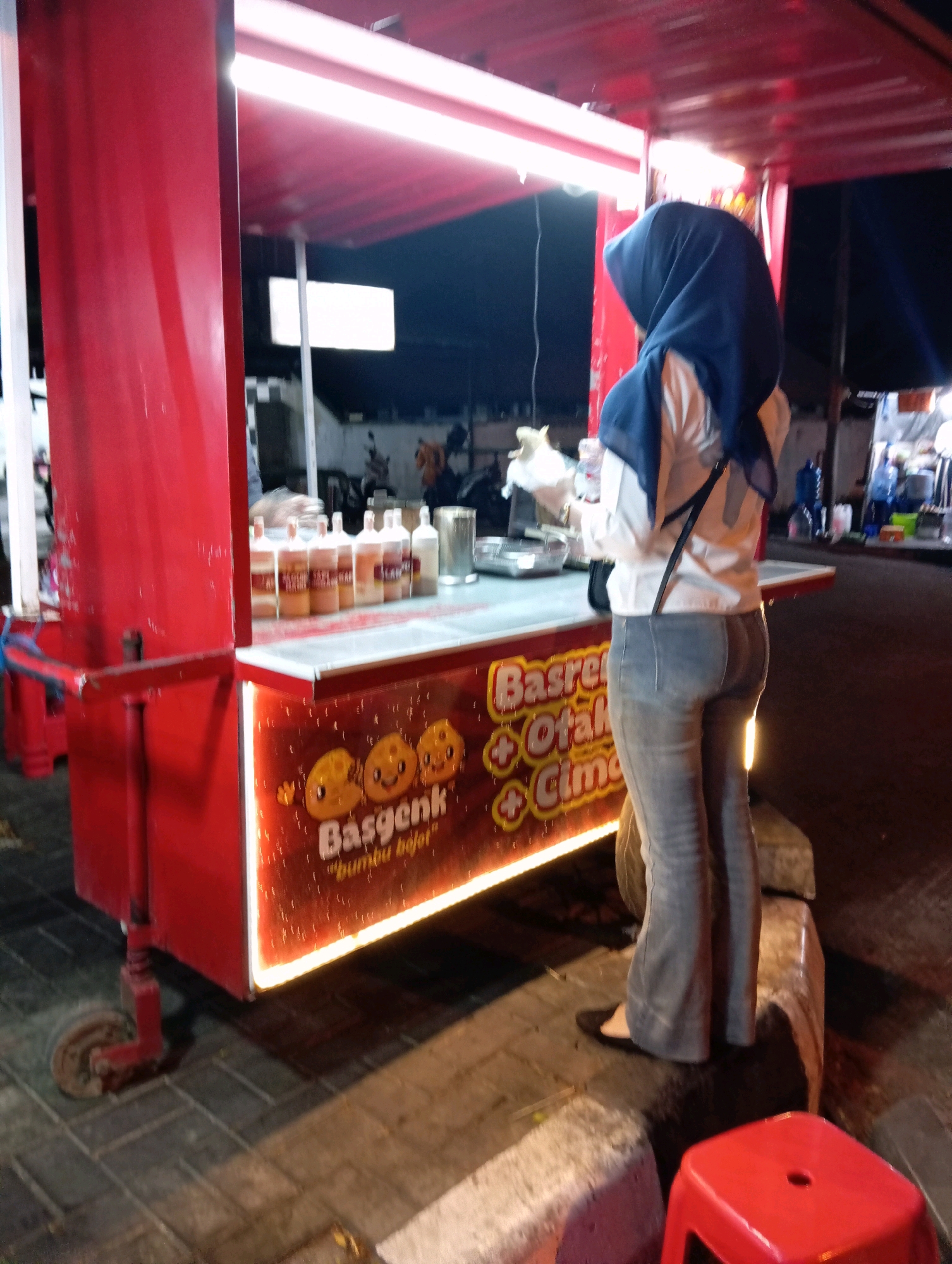 A woman wearing a blue hijab, white shirt, and grey flared jeans stands behind a red food cart. The cart has a counter displaying several bottles and a lit sign with text and cartoon images of happy faces. The background shows a street scene at night with other stalls and a red stool in the foreground. The food cart appears to be located on a tiled sidewalk in Magelang, Indonesia.