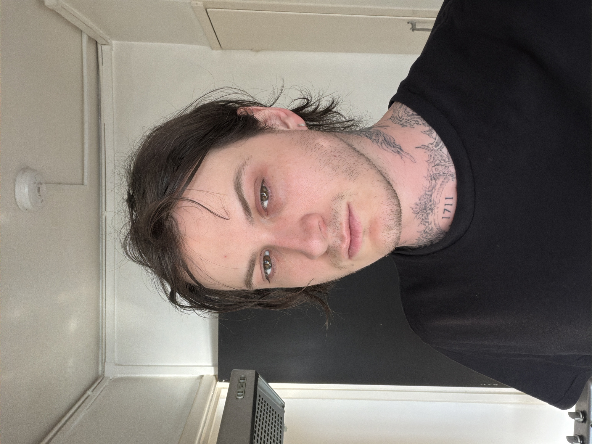 A light-skinned individual with brown hair, facial hair, and tattoos on their neck is wearing a black t-shirt. They are indoors in a room with white walls.