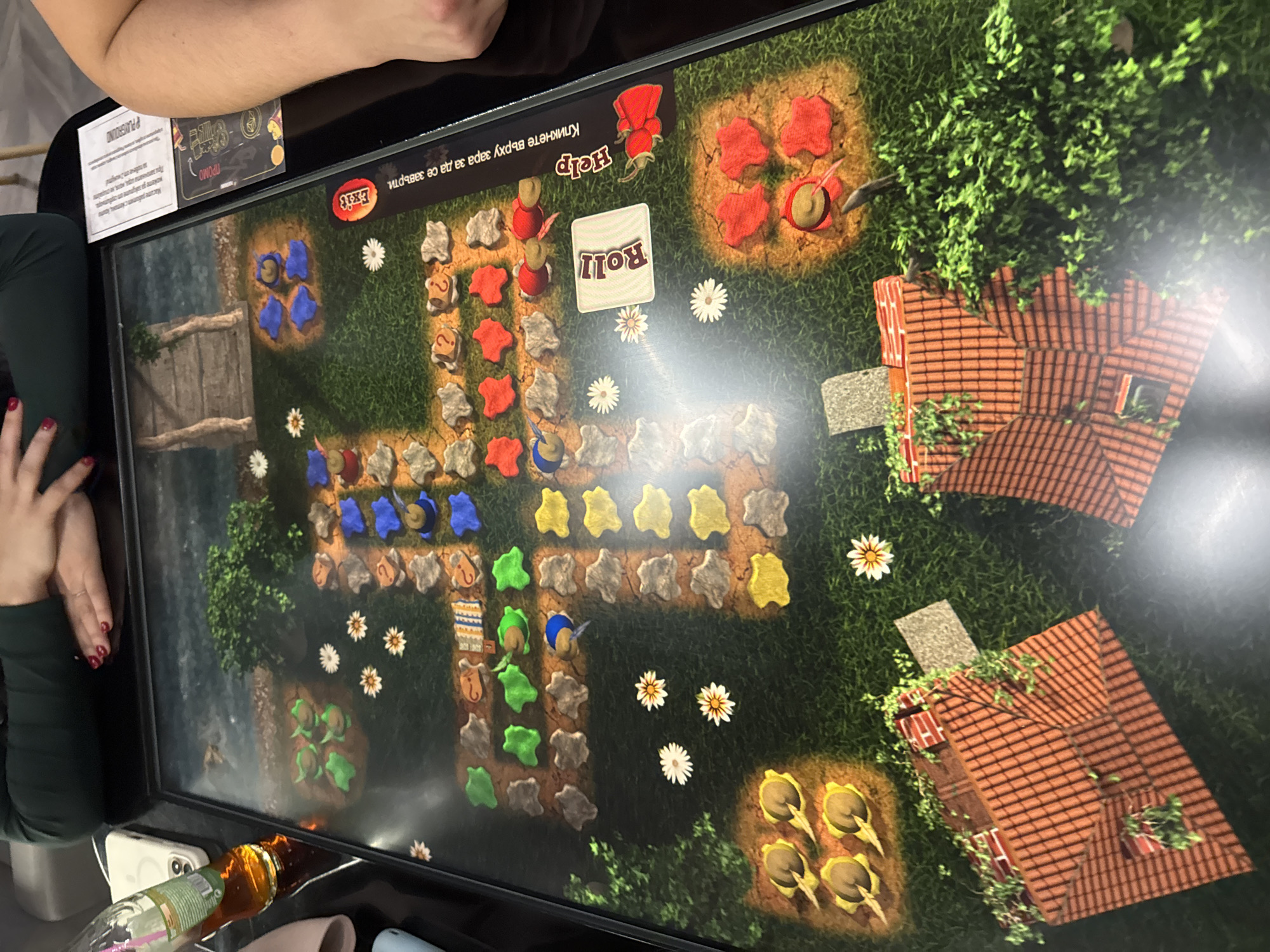 The image shows a screen displaying a digital board game, likely situated on a table within an indoor setting. The game features a path composed of variously colored spaces (blue, red, yellow, green) with flower accents leading to houses and other features. A "Roll" button is visible in the game's interface. Parts of two individuals are also in the image. One on the top left, with their arm visible and another on the left side, with part of the person's legs and hands with red painted fingernails. Additionally, a bottle of liquid, a phone, and other personal items lie near the screen. There is no information within the image or the OCR text that allows for the location to be determined to be in Sofia, Bulgaria.