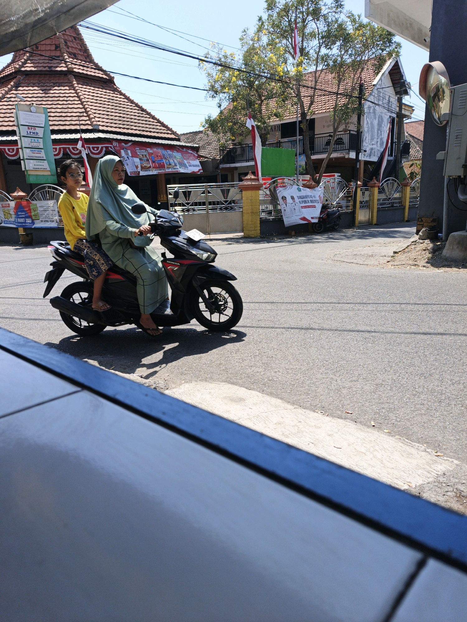 In Ketegan, Indonesia, on a sunny day, a woman wearing a light green hijab and a woman with glasses wearing a yellow shirt are riding a black Honda motorcycle on a paved road. The woman in front is operating the motorcycle, while the woman behind her is seated. The background shows buildings with red tile roofs, some adorned with Indonesian flags and banners. A tree with yellow flowers is visible, and utility lines stretch across the sky. The scene is observed from a low angle, with a dark blue object in the foreground partially obscuring the view.