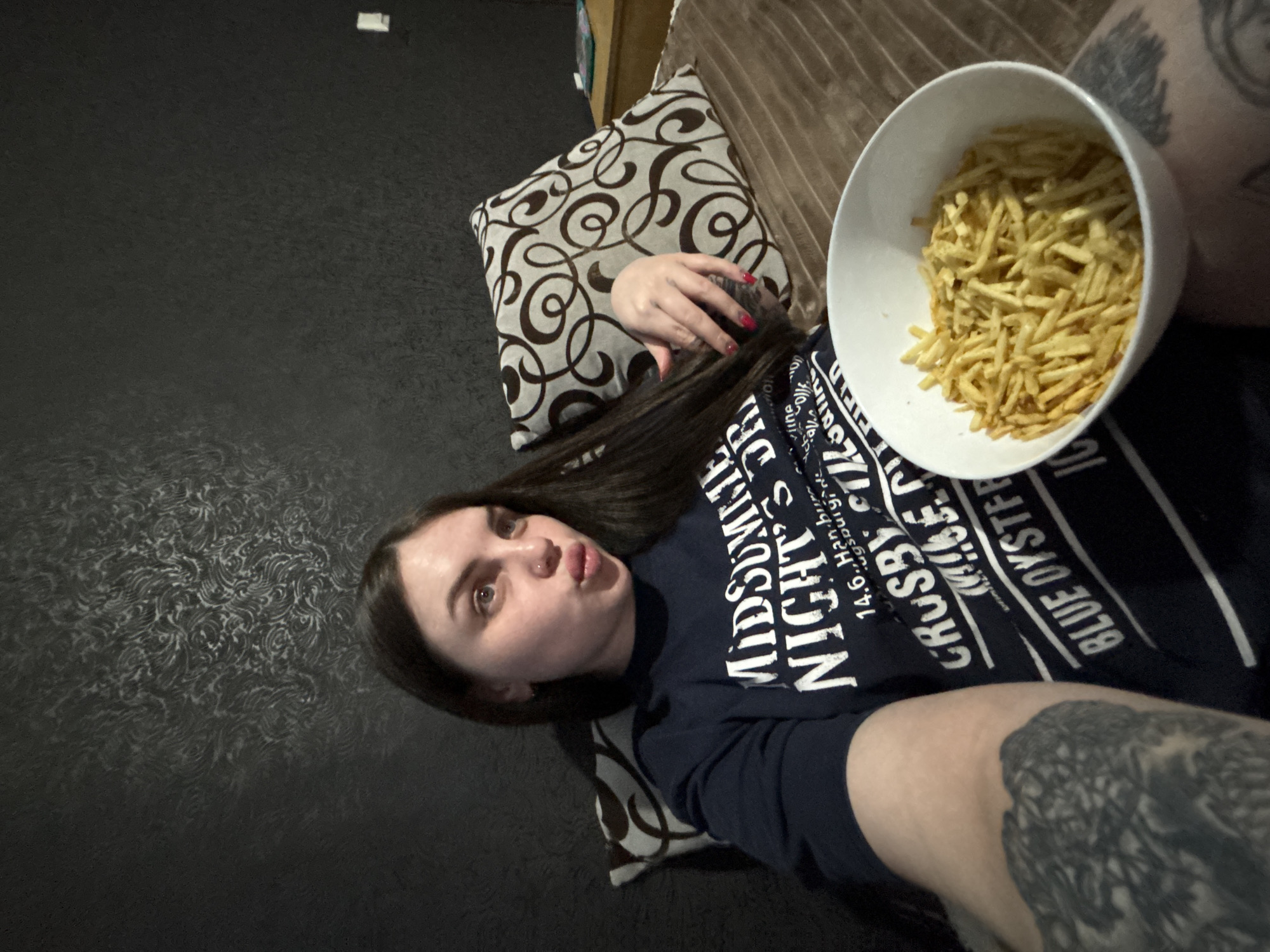 A woman lies on a bed or sofa with a decorative pillow behind her head. She is wearing a dark blue t-shirt with white text. She holds a large white bowl filled with shoestring potatoes or fries. The woman has dark hair and pale skin, and is making a slightly puckered facial expression. Her fingernails are painted red. Part of a tattoo is visible on her arm, near the bowl. The background consists of a patterned wall on one side and a brown, possibly upholstered, surface on the other. The location in the city of Chornomorskoe, Ukraine, is not discernible.