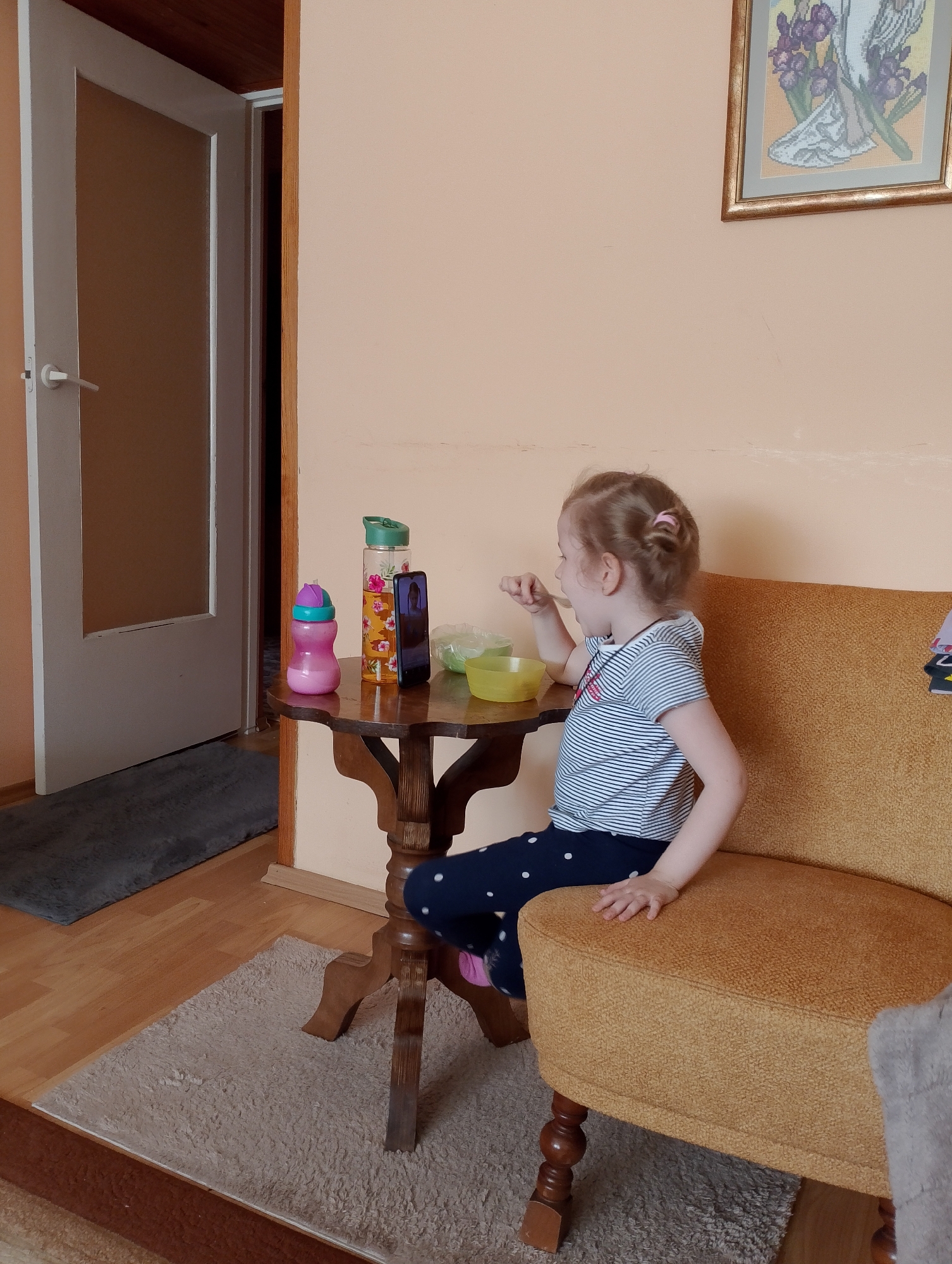 In this indoor shot, a young girl with blonde hair is sitting on an orange upholstered chair and eating. She is wearing a blue and white striped t-shirt and dark blue pants with white polka dots. Her hair is pulled back with a pink hair tie. She is looking to her left and eating from a yellow bowl with a spoon. In front of her is a small, wooden table with a phone and two water bottles on it. One water bottle is clear with a floral pattern and a green lid, while the other is pink with a purple lid. The girl is on the right side of the image, and the table is in the center.
Behind the girl, the wall is painted a peachy orange color. A framed cross-stitch picture of an angel with purple flowers hangs on the wall above and to the right of the girl. To the left of the girl is a doorway with a white door that is partially open, revealing a dark interior. A dark gray rug is on the floor in front of the doorway. A lighter colored shaggy rug is on the wooden floor in front of the chair and table. The lighting suggests it is daytime, but there are no windows visible to indicate the time of day or weather. The scene appears to be in a home.