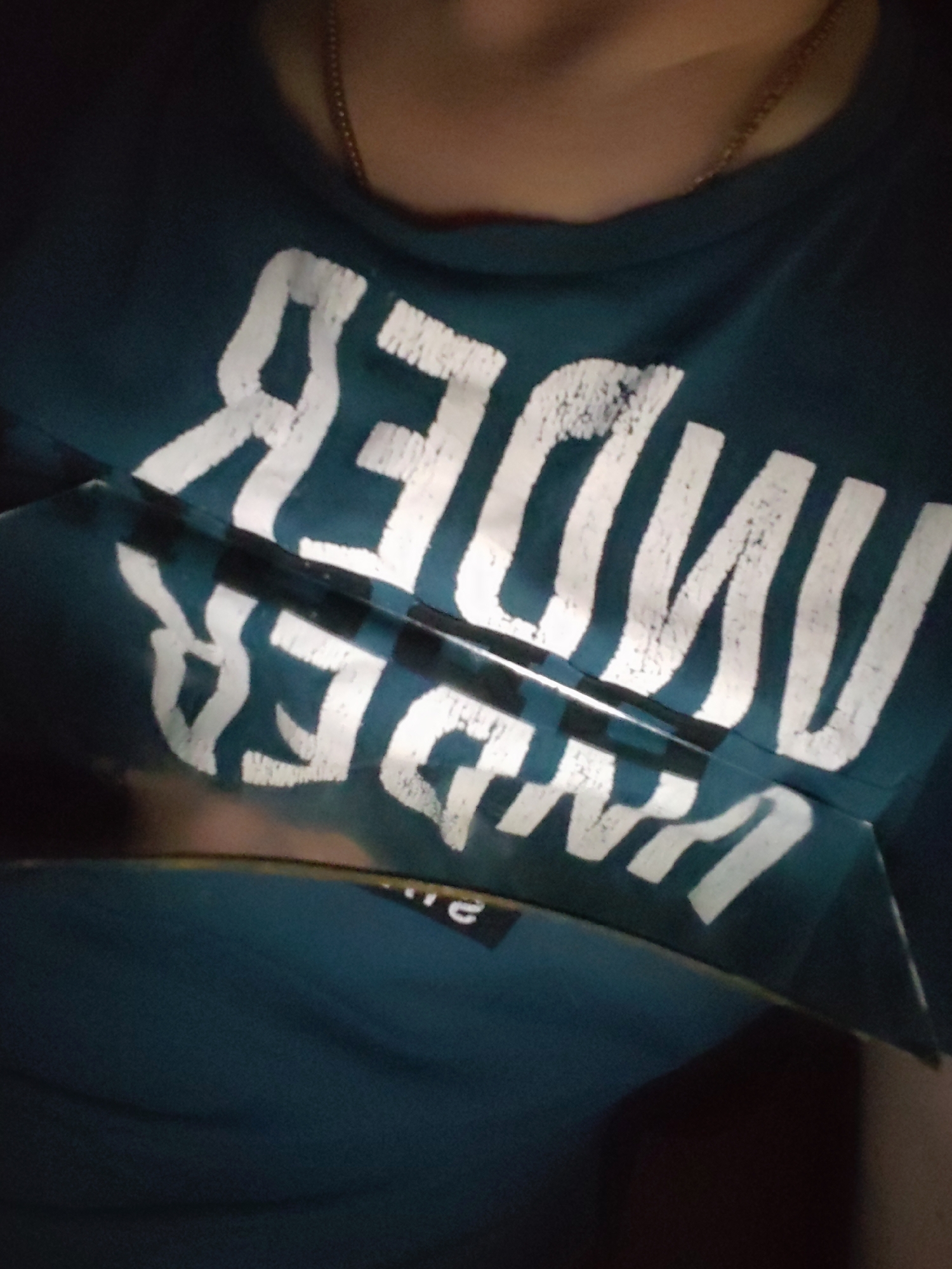 A close-up shot shows a person wearing a teal t-shirt with the word "UNDER" printed in white letters. A reflective object, possibly a piece of glass, is placed over the shirt, creating a mirrored and distorted reflection of the word. A gold chain is visible around the person's neck. The environment appears dark, with limited lighting. No specific location in Chaikovsky, Russia can be determined from the visual information provided.