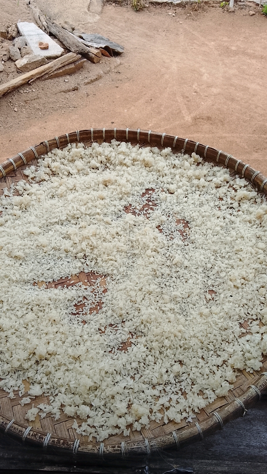 In Way Melan, Indonesia, a woven bamboo tray filled with uncooked white rice sits on a wooden surface. The tray is round with a rim of tightly bound twine. The rice is spread evenly across the tray, with some areas showing the woven pattern of the bamboo underneath. In the background, the ground is a reddish-brown dirt, and scattered debris, including pieces of wood and stone, are visible. The scene appears to be an outdoor setting.