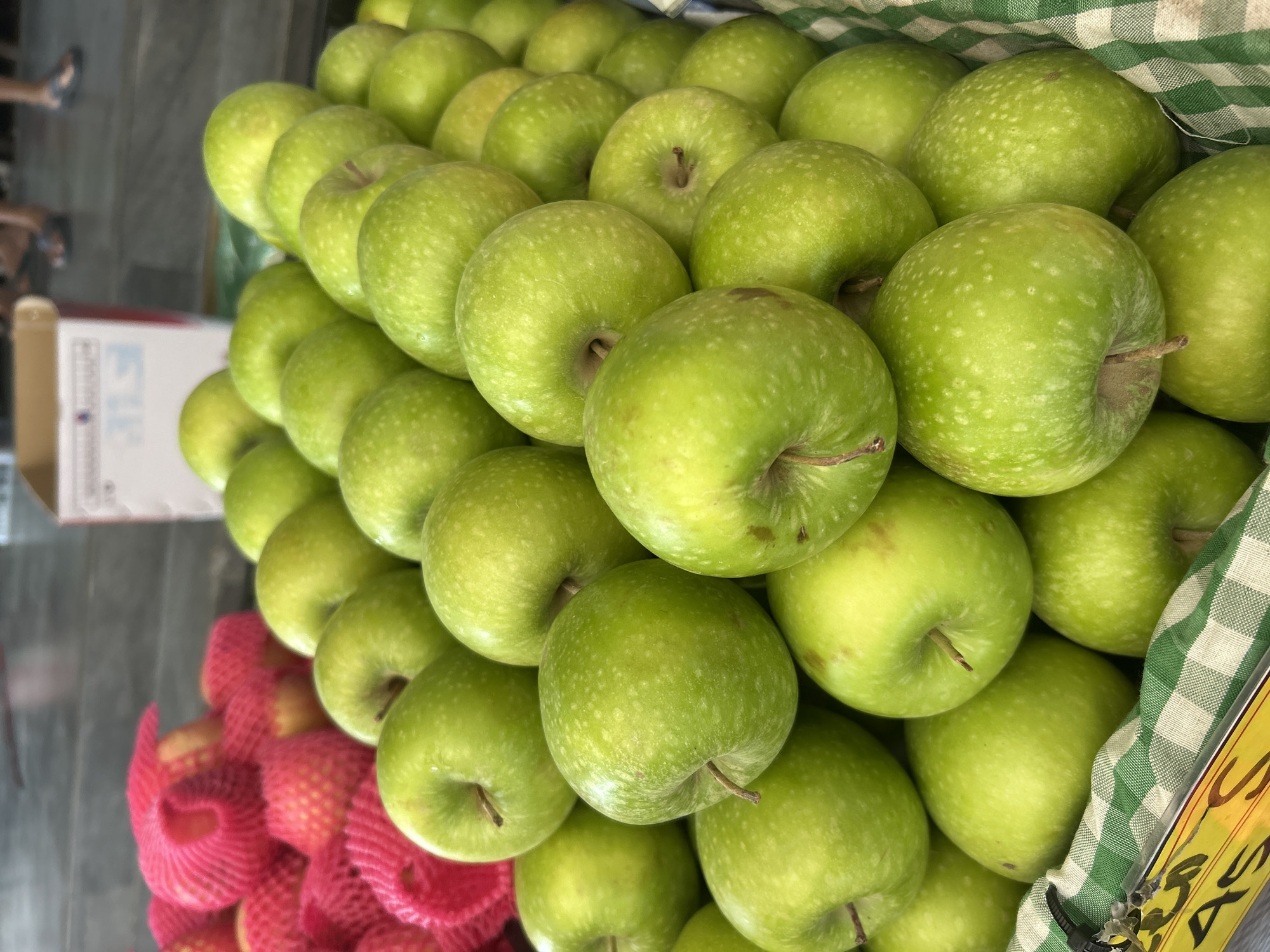 A close-up, eye-level shot shows a large display of green apples at a market in Sukawati, Indonesia. The apples are piled high in a basket lined with a green and white checkered cloth. The apples are mostly round with some slight imperfections and small white speckles. In the background, out of focus, are several red apples encased in pink netting, and a portion of a white cardboard box. The floor is visible in the background and appears to be made of concrete. Two people are partially visible in the far background, their legs and feet only showing.