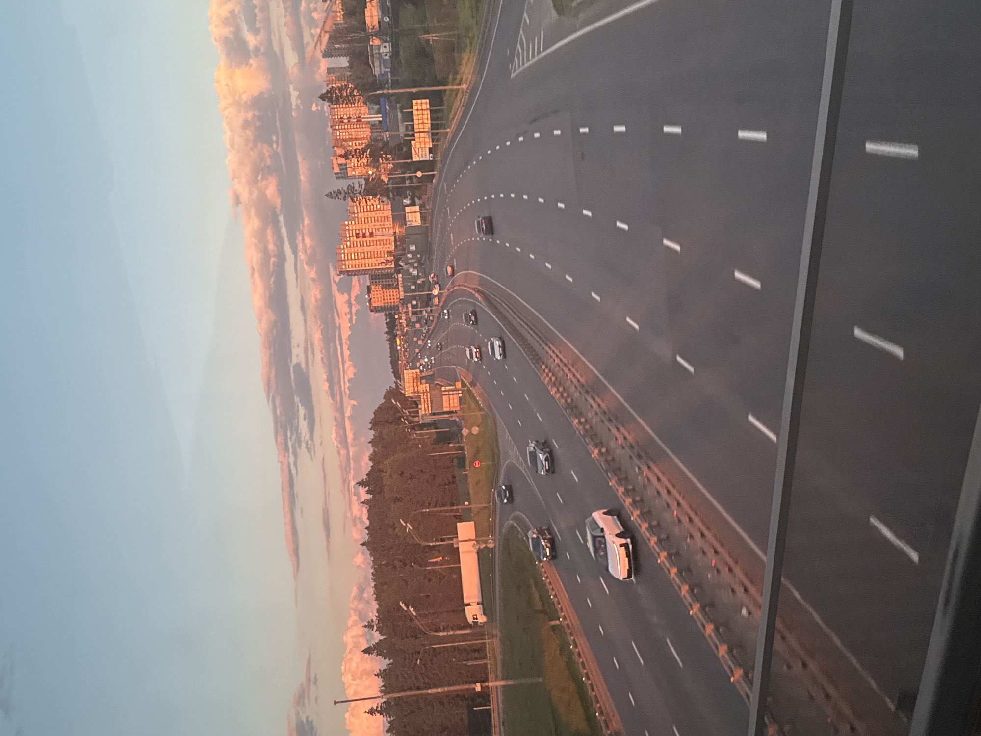 The image, rotated 90 degrees counter-clockwise, presents an elevated view of a multi-lane highway or motorway during what appears to be sunset or sunrise. The scene captures a moment during the "golden hour," bathing the landscape in warm, orange-pink light.
The primary subject is the expansive highway, curving gently through the frame. Multiple vehicles, including cars and at least one truck, are visible on the road, indicating active traffic flow in both directions. Lane markings, consisting of dashed white lines, are clearly visible, as are solid white lines and a concrete barrier separating the main thoroughfare. A red circular "No Entry" sign is visible on the median strip.
To the left of the highway, a series of multi-story residential buildings with orange facades are illuminated by the low sun, creating a striking contrast with the blue sky above. Below these buildings, trees and streetlights line the road. To the right of the highway, a dense forest or wooded area runs parallel to the road.
The sky above is a notable feature, displaying a gradient from a clear, soft blue at the top to vibrant orange and pink hues where scattered clouds catch the last or first rays of the sun. The perspective is from an elevated vantage point, likely a bridge or overpass, with a part of its structure visible in the foreground. No people are discernible in the image. The overall impression is one of calm and tranquility, despite the movement of traffic. The location context is посёлок опытного хозяйства «Ермолино», Russia.