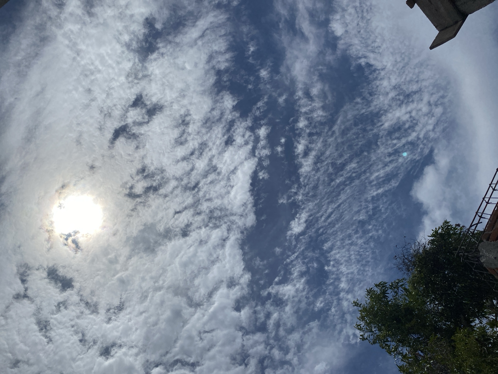 This is a low-angle, full-frame shot of a cloudy sky with the sun visible in the upper center of the frame. Wispy, white clouds are scattered across a deep blue sky, creating a textured pattern. The sun is partially obscured by clouds, casting a bright glow that illuminates the surrounding cloud formations. In the lower left corner, the dark green foliage of a tree is visible, extending upwards into the sky. A partially constructed building or structure, made of concrete and brick with some exposed rebar, occupies the lower right portion of the image. The overall impression is one of a bright, slightly overcast day, viewed from a ground-level perspective.