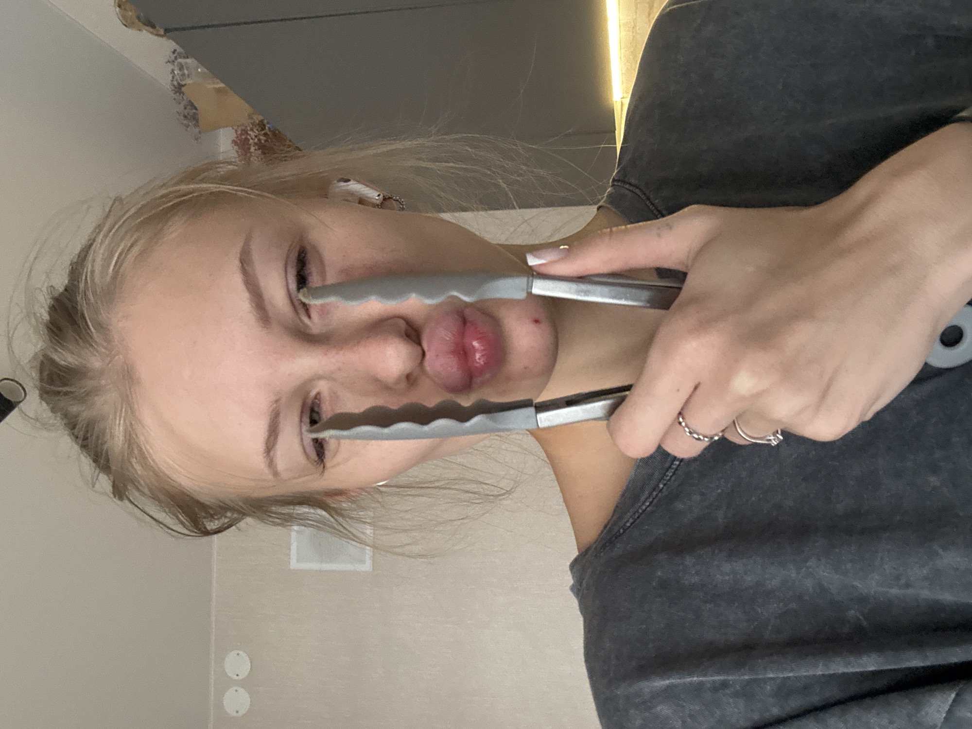 A young Caucasian woman with blonde hair pulled back is holding two metal kitchen tongs in front of her face, one on each side of her nose, with the tips touching her cheeks near her eyes. She is looking directly at the camera with her lips pursed, creating a comical expression. She is wearing a dark gray t-shirt and several rings on her fingers. There are white wireless earbuds in her ears. The background appears to be an indoor setting, possibly a kitchen, with a light-colored wall and cabinetry visible. The lighting suggests it is daytime. There is no indication of the city of Perm, Russia in the image.