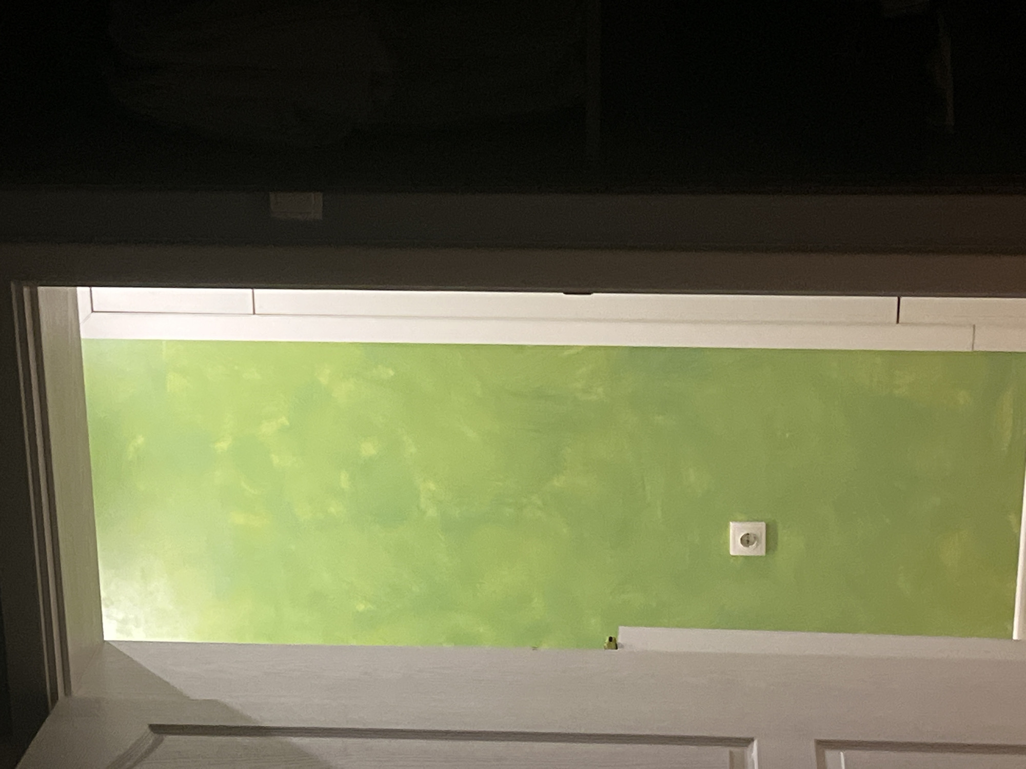 A low-angle shot framed by a white door and its upper frame, reveals a green wall with a textured pattern. A white European-style electrical outlet is visible on the wall. The upper portion of the image is dominated by a dark, out-of-focus area. The location is inside a building in Pokrovnik, Bulgaria.